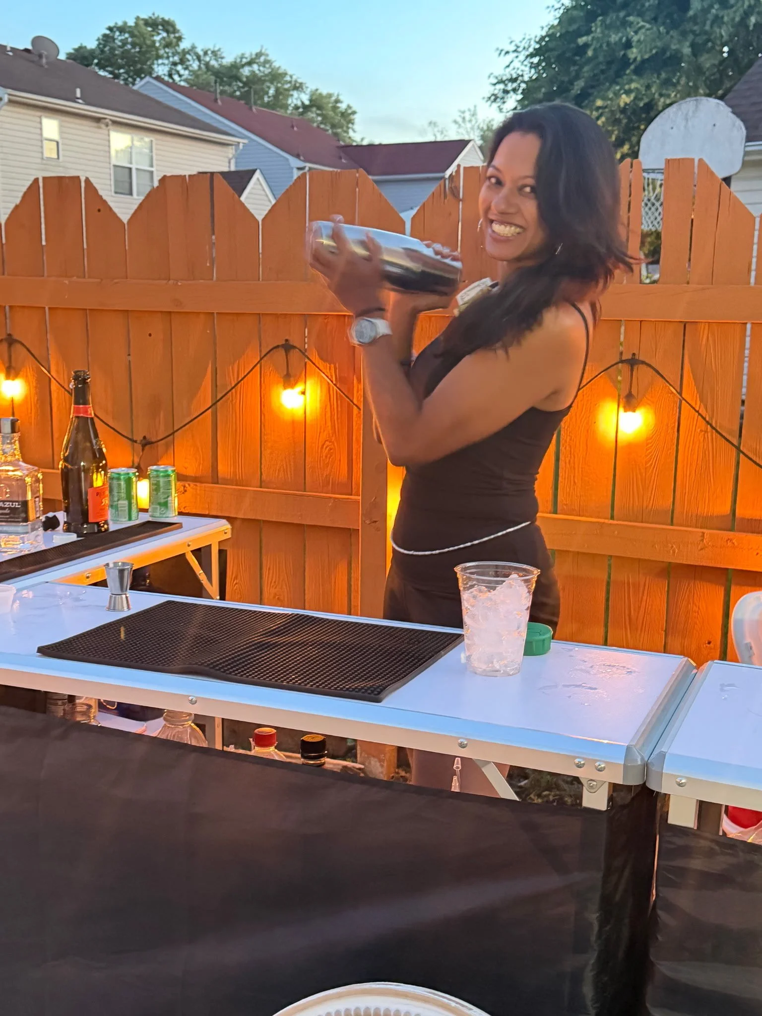 A smiling bartender shaking and holding a metal cocktail Boston shaker at an outdoor backyard party. There are bottles, cans, and a plastic cup on a table, with string lights on a wooden fence in the background.