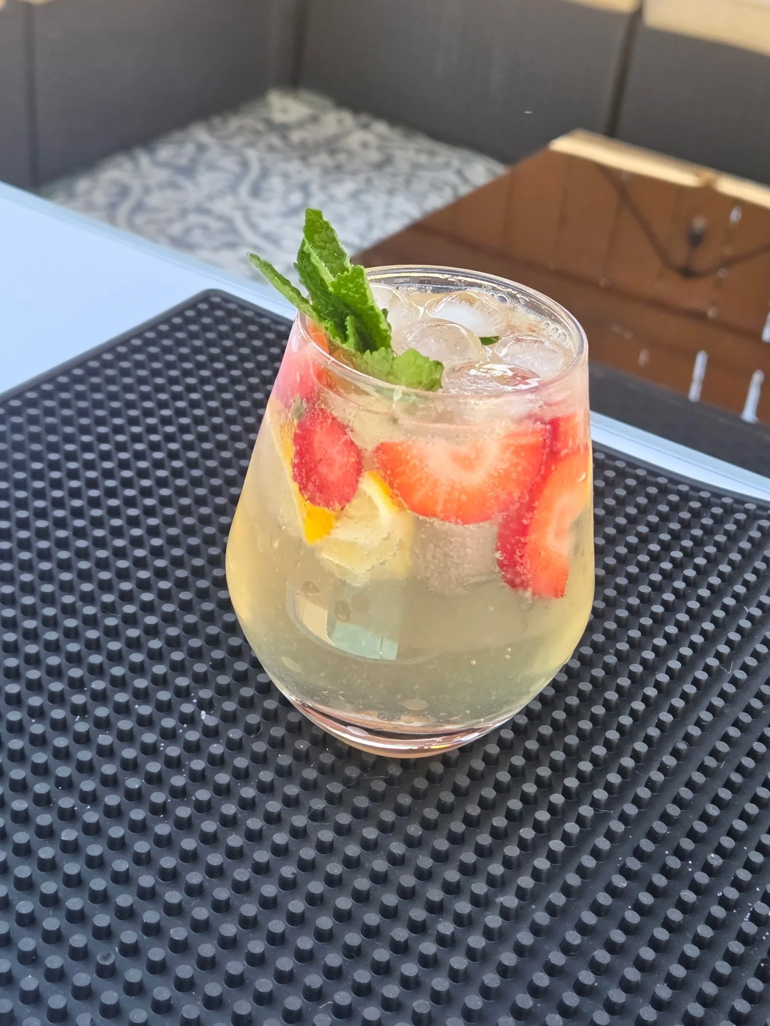 A glass of lemonade spritz with slices of lemon and strawberries, ice cubes, and a sprig of mint on top, placed on a black textured surface which is a bar mat..