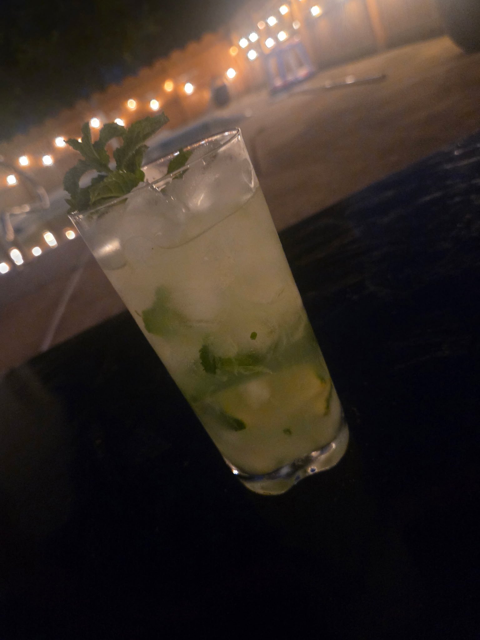 A cocktail drink which could be a mojito with ice, mint leaves, and lime slices in a glass, against a background of string lights at night.