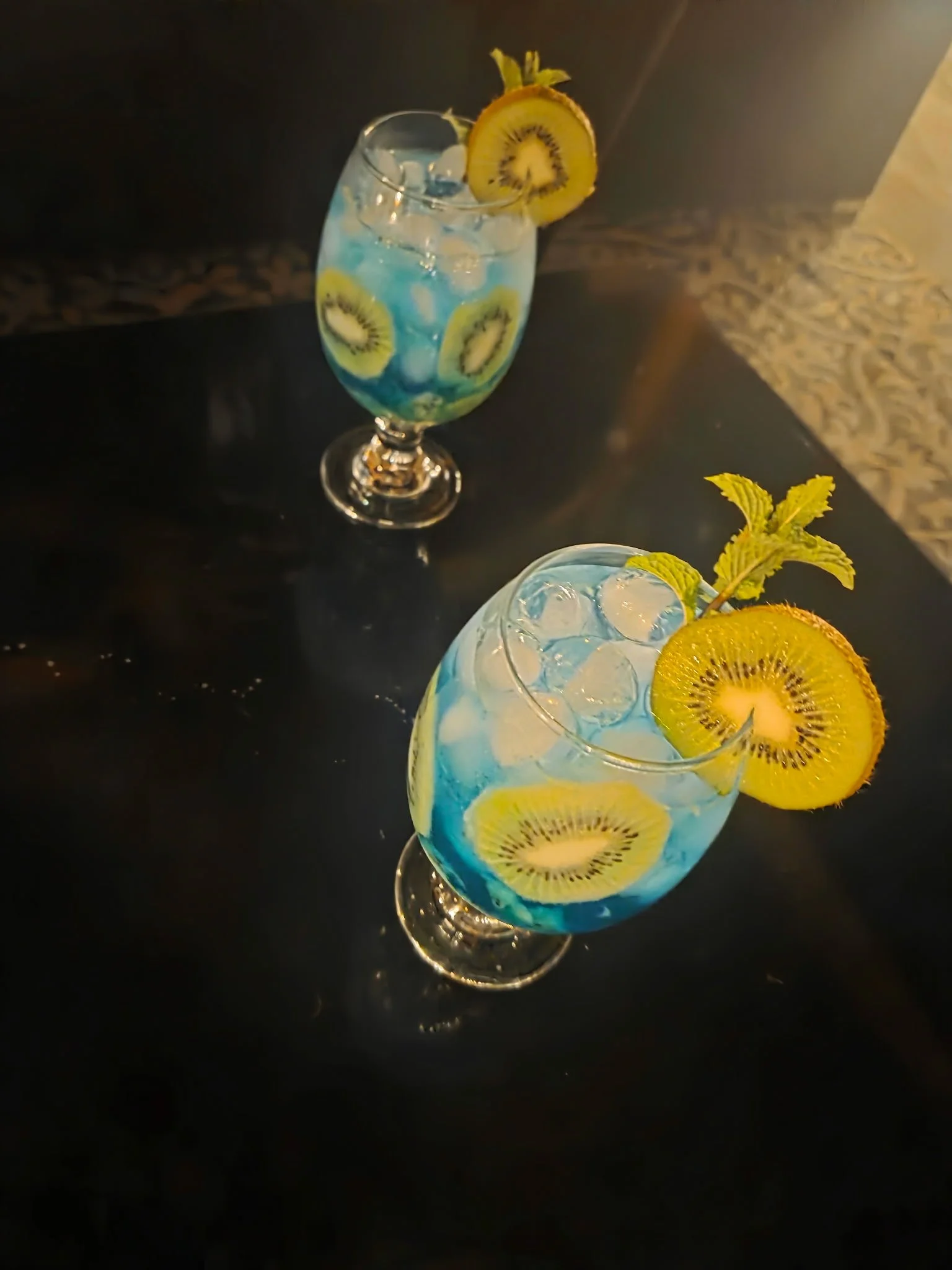 Two tropical drinks with ice, decorated with slices of kiwi and lemon, garnished with mint leaves, on a dark surface.