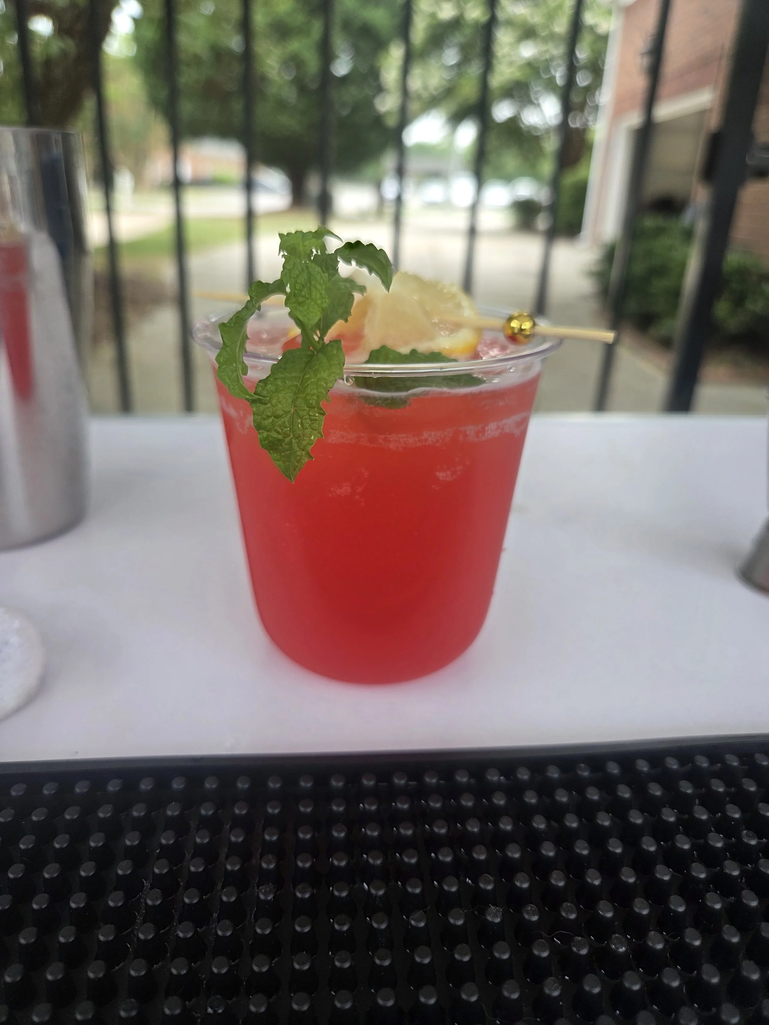A red cocktail drink garnished with a sprig of mint, lemon wedge, and a cocktail pick with a small yellow decorative bead, placed on a white surface in front of a blind or window with trees outside.
