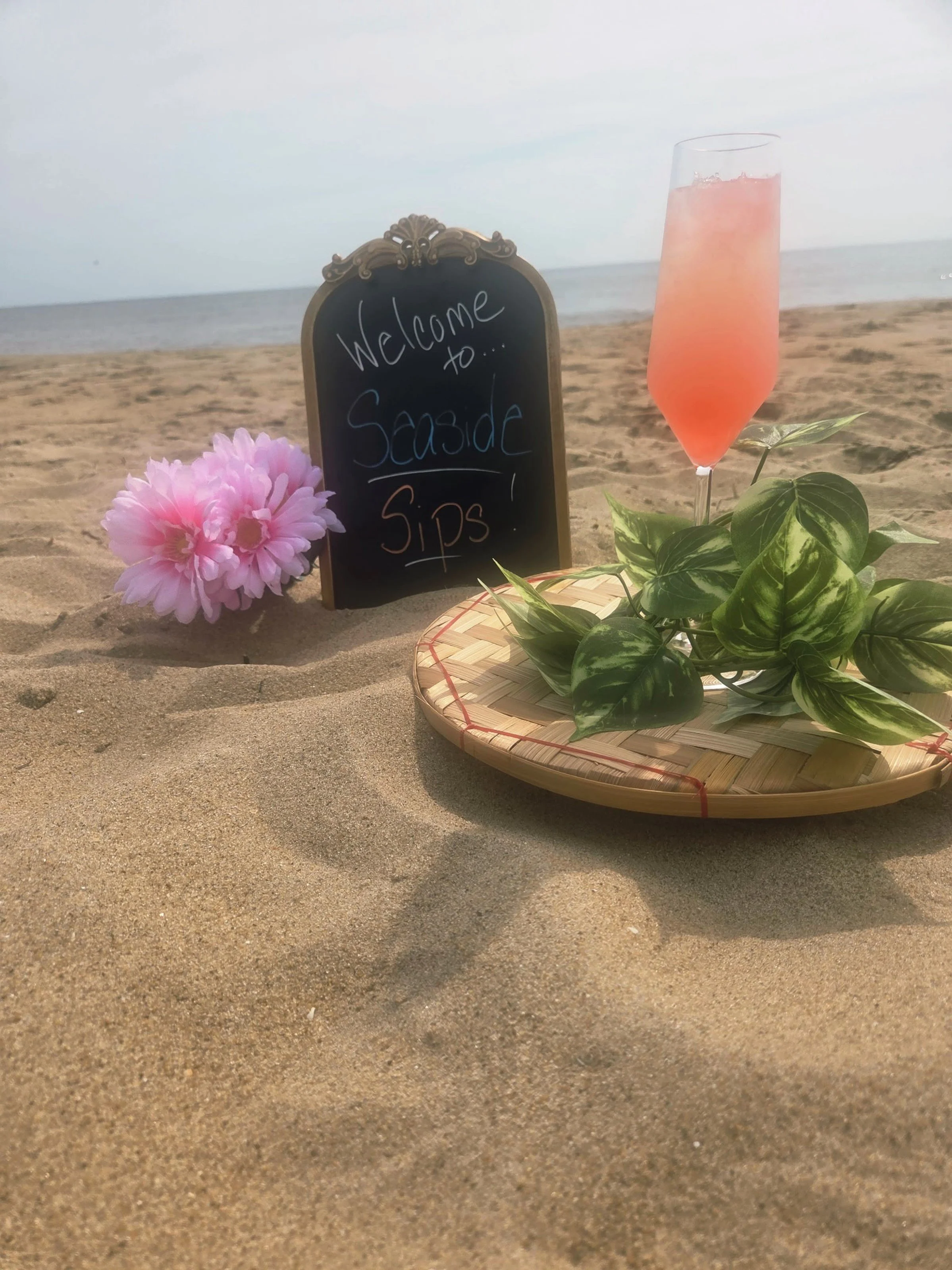 A beautiful pink mocktail with pathos leaves around the base on a wooden plate placed on   a sandy beach with a sign behind it reading "Welcome to Seaside Sips" and pink flowers next to it and in front of the ocean.