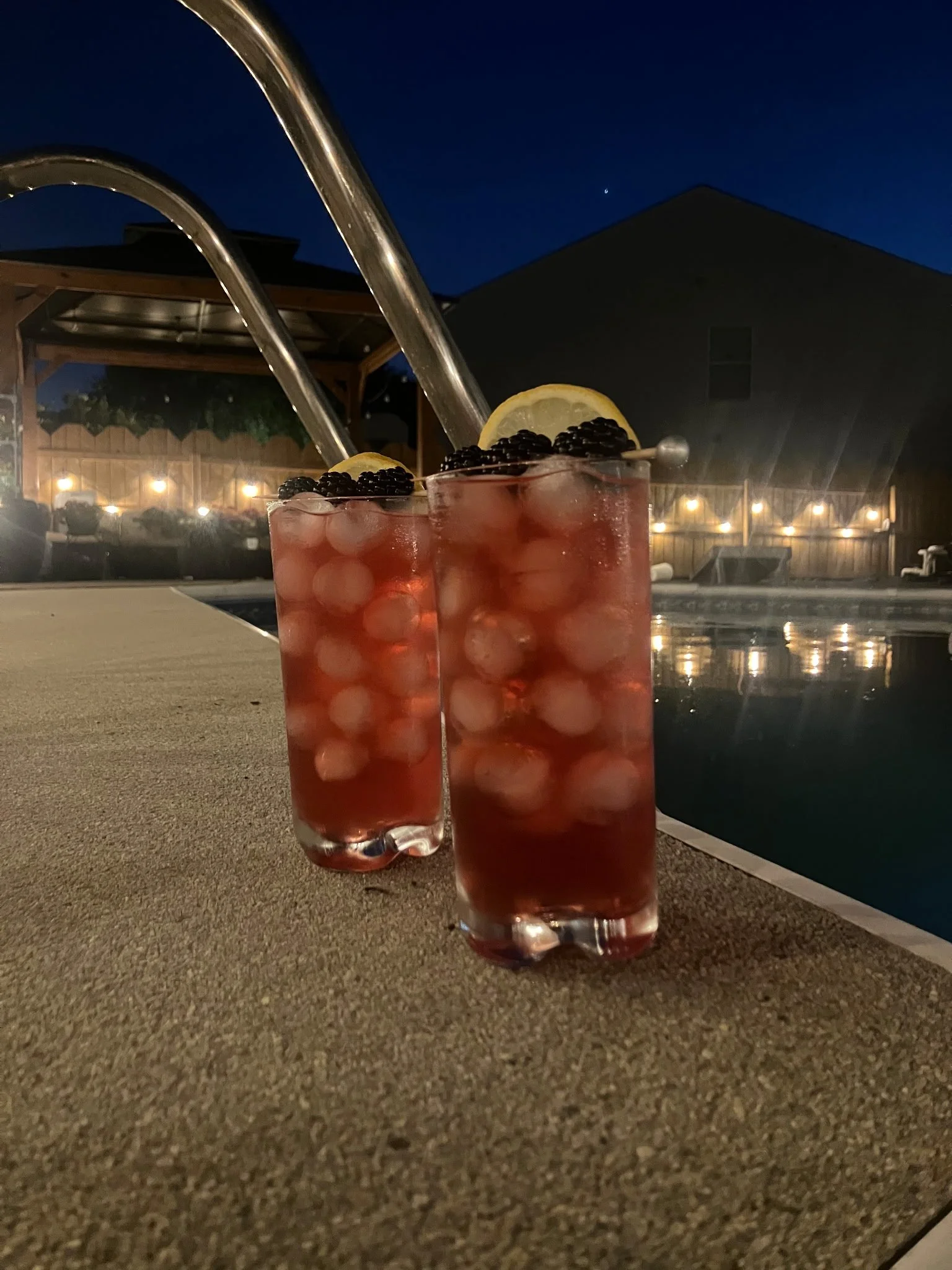 Two glasses of pink-colored beverage named "Neptune's Kiss" with lemon slices and blackberries, placed on a concrete surface near a swimming pool at night, with a house and string lights in the background.