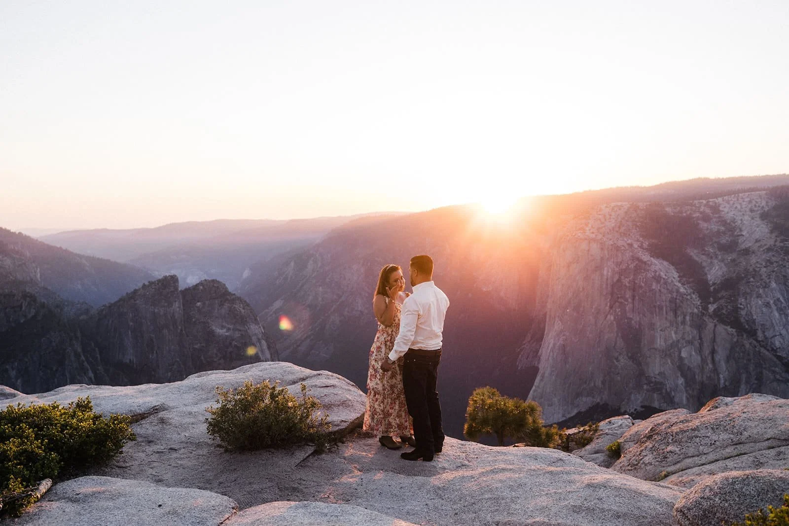 Couple standing on a rocky ledge at Taft Point in Yosemite during their engagement session, sun low on the horizon casting a warm glow