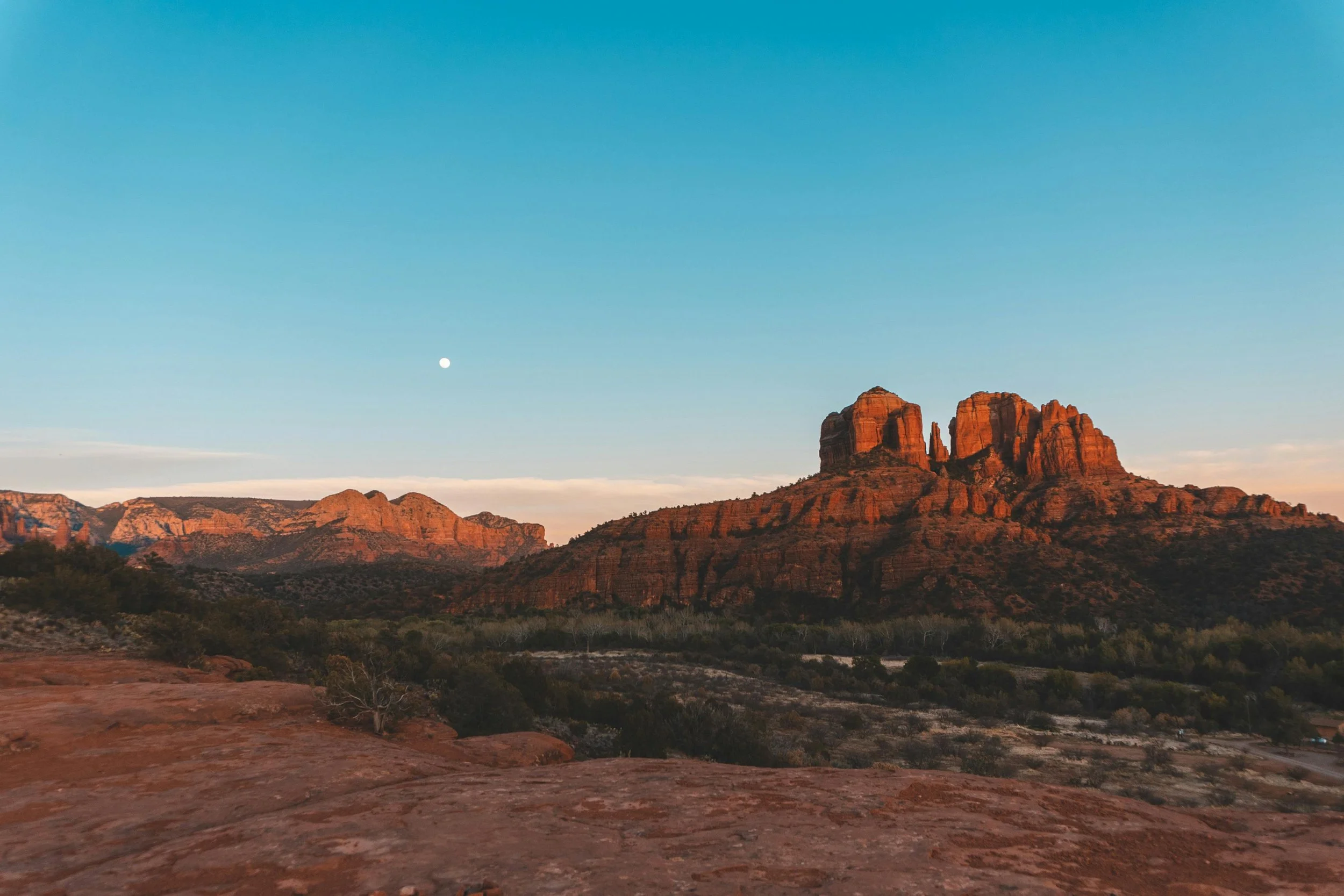 View of red rock formations in a desert landscape under a clear blue sky with the moon visible near the horizon.