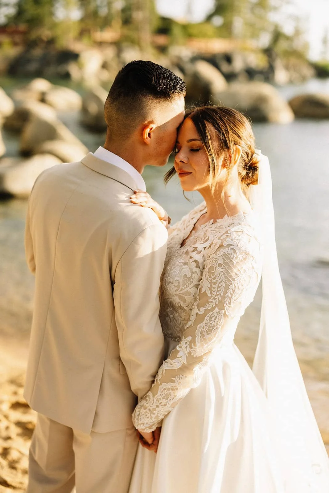A couple in wedding attire embracing on a rocky beach during sunset.