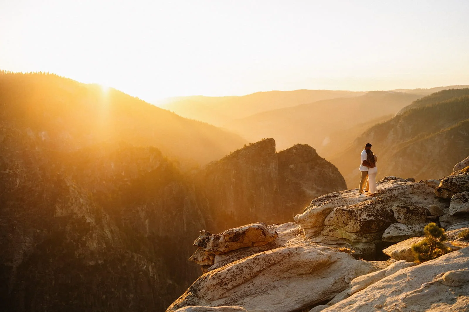 Couple hugging on the cliffs of Taft Point in Yosemite at sunset during their engagement session, canyon and mountains behind them