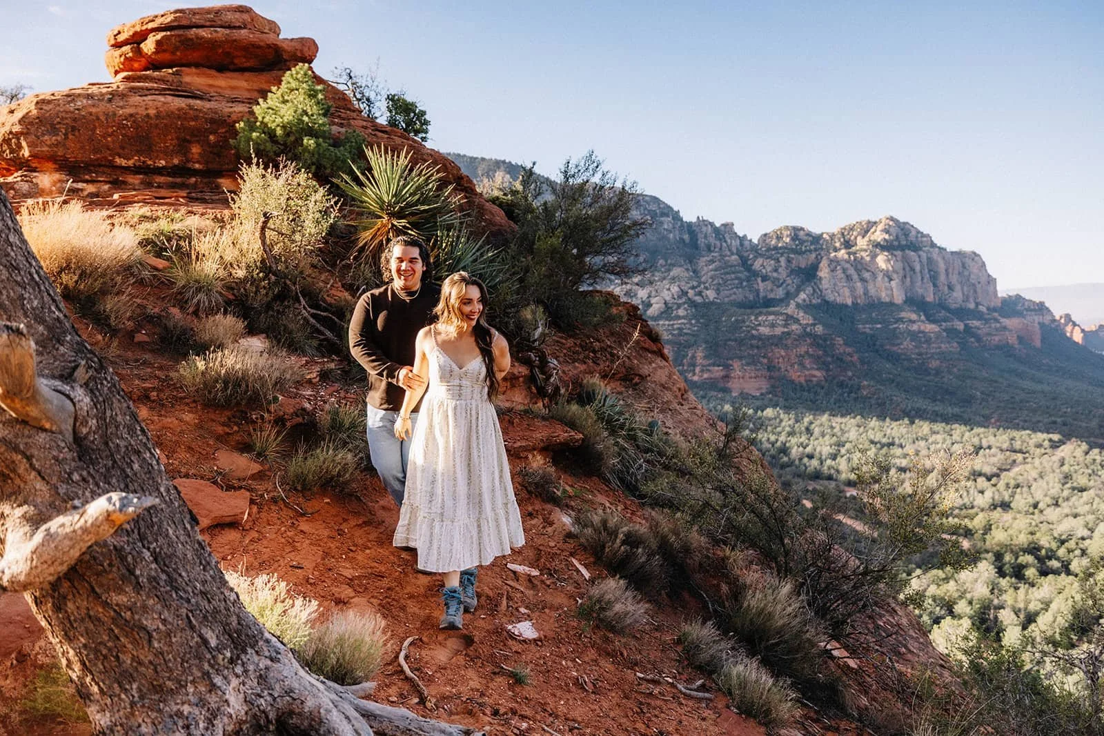Couple hiking through Sedona's red rock landscape during their engagement session, green desert plants and mountains around them