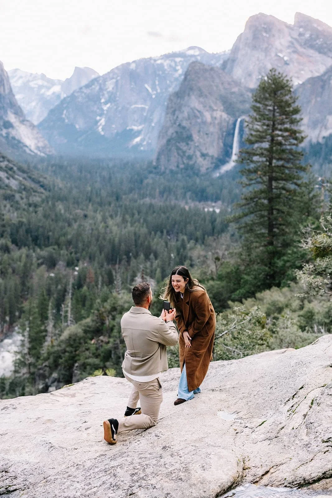Man kneeling with a ring box in front of his partner at Tunnel View in Yosemite, mountains, trees, and a waterfall behind them