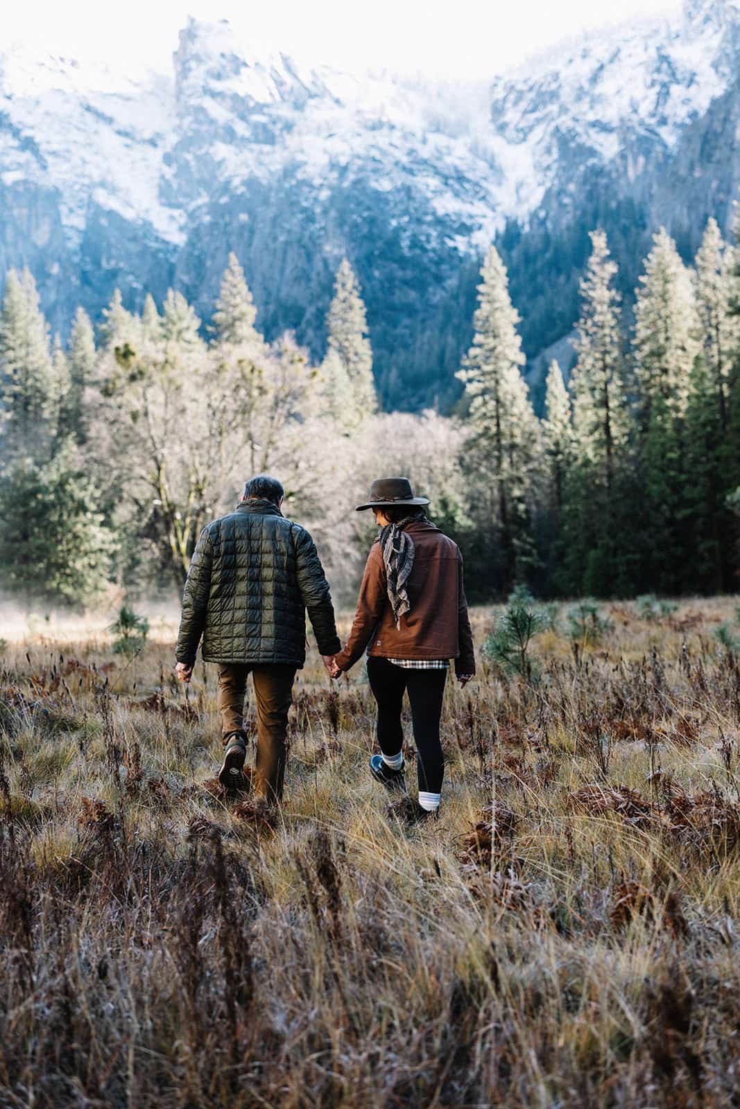 Couple walking hand in hand through Cathedral Meadow in Yosemite, snow-capped mountains and evergreen trees behind them