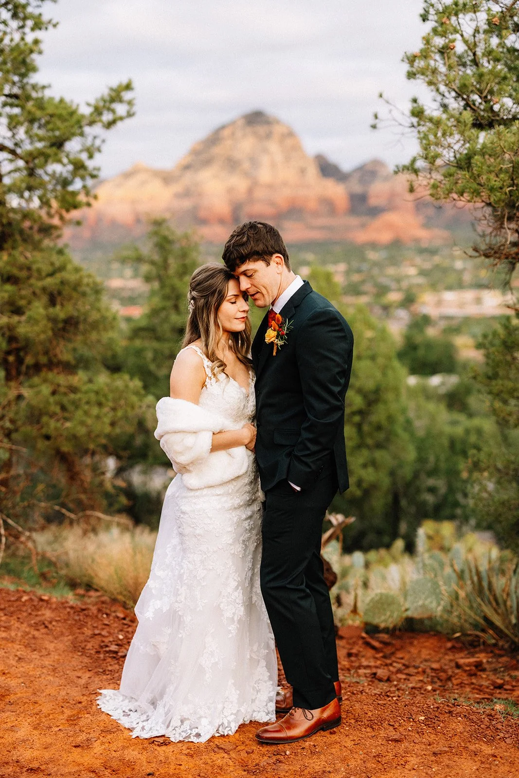 A bride and groom standing close with foreheads touching outdoors, with red rock formations and desert vegetation in the background.