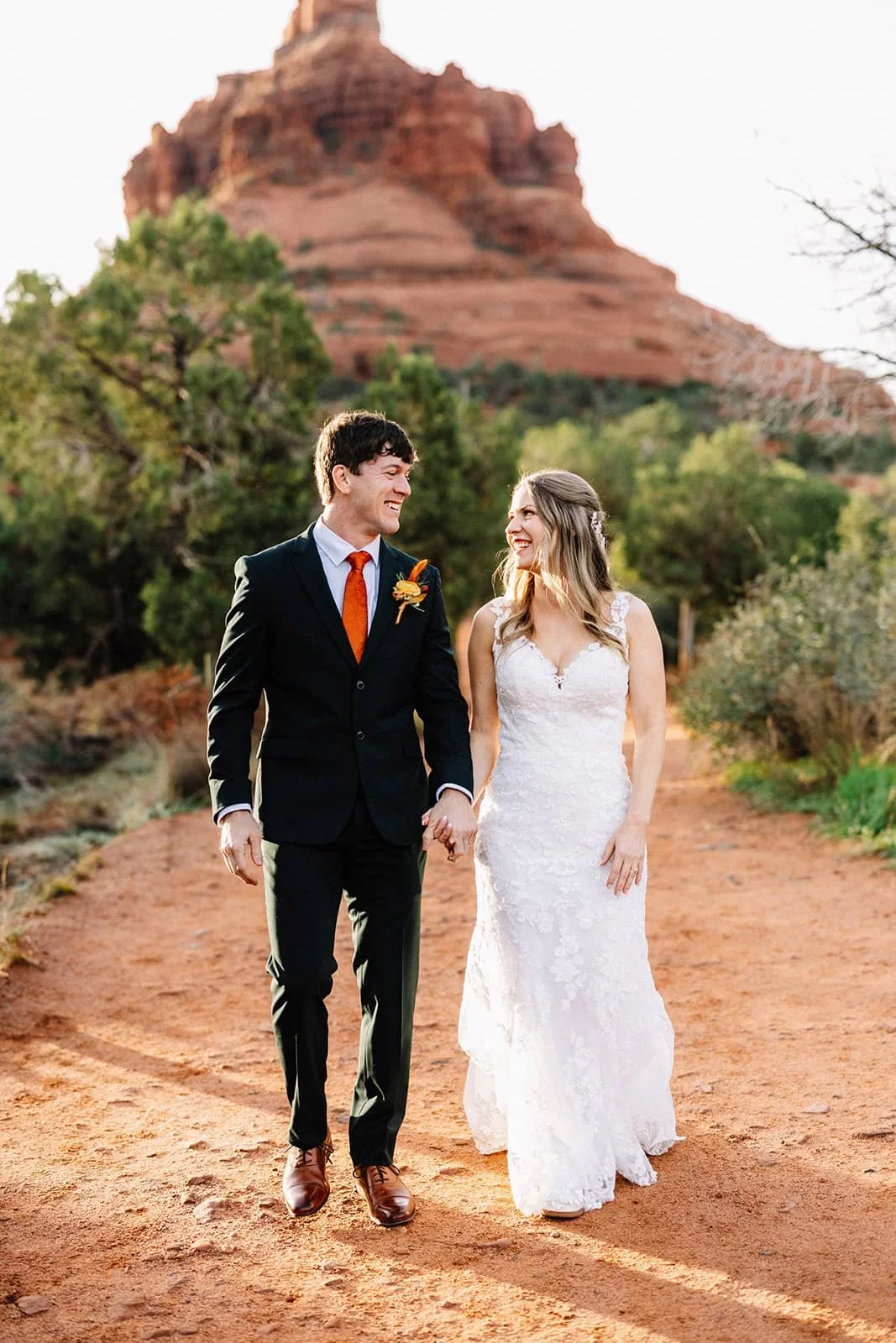 Bride and groom holding hands as they walk a dirt path through Sedona's red rock formations