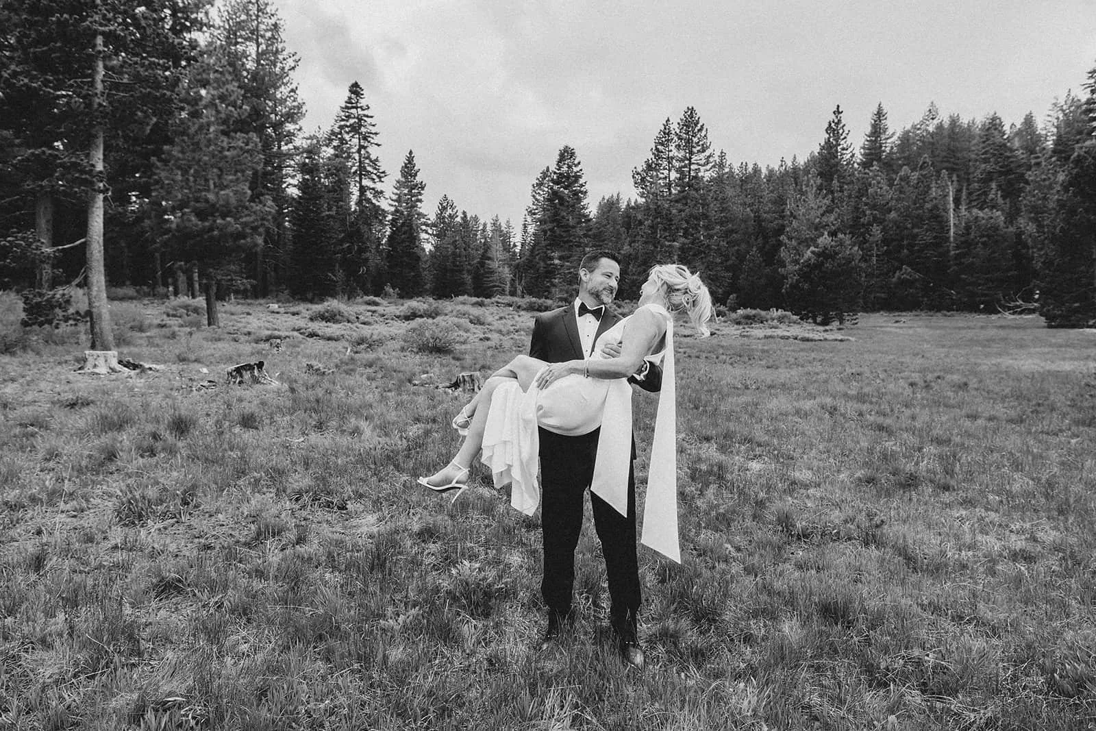 Groom carrying the bride through an open meadow surrounded by pine trees.jpg