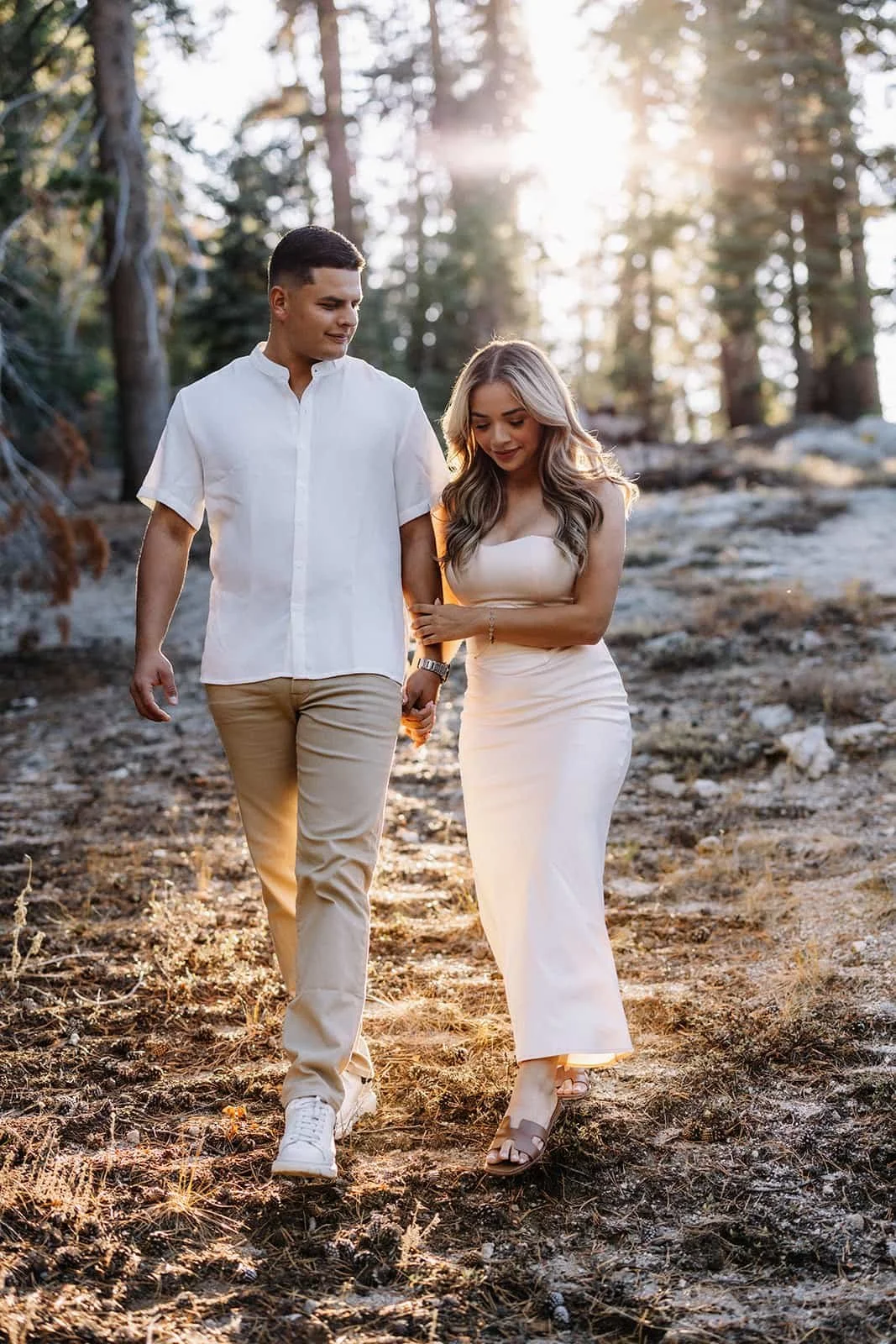 couple walking hand in hand through the woods at Taft Point in Yosemite at sunset, dressed in light-colored clothing
