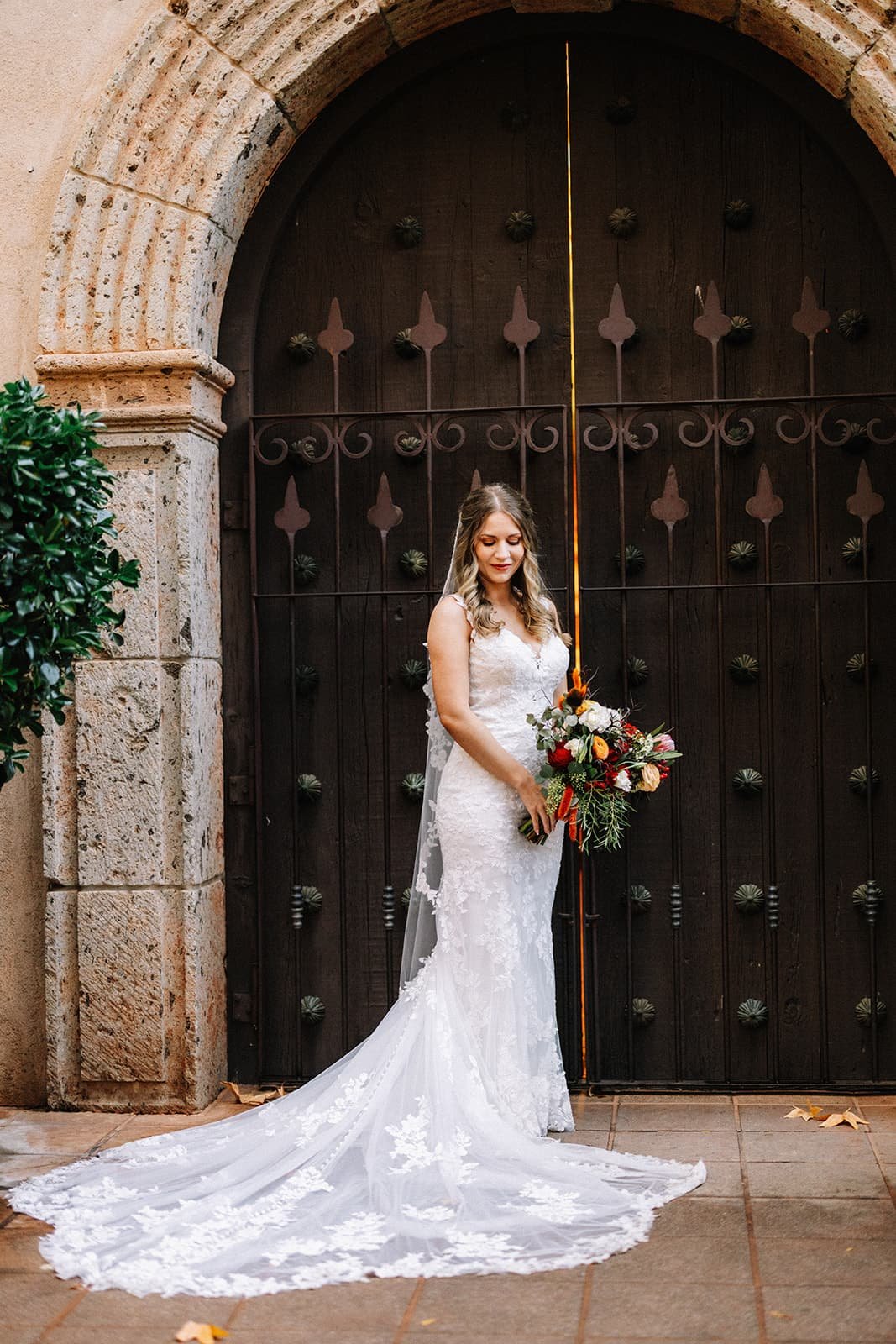Bride in a white lace gown holding a vibrant bouquet in front of a dark wooden door with ironwork detail during her Sedona elopement