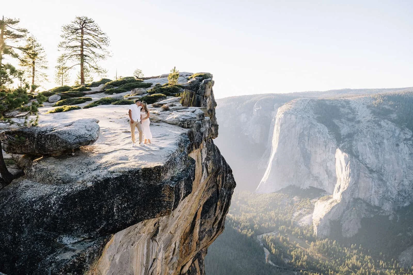 Couple standing on a cliff edge at Taft Point in Yosemite, overlooking the canyon with a distant waterfall during their engagement session