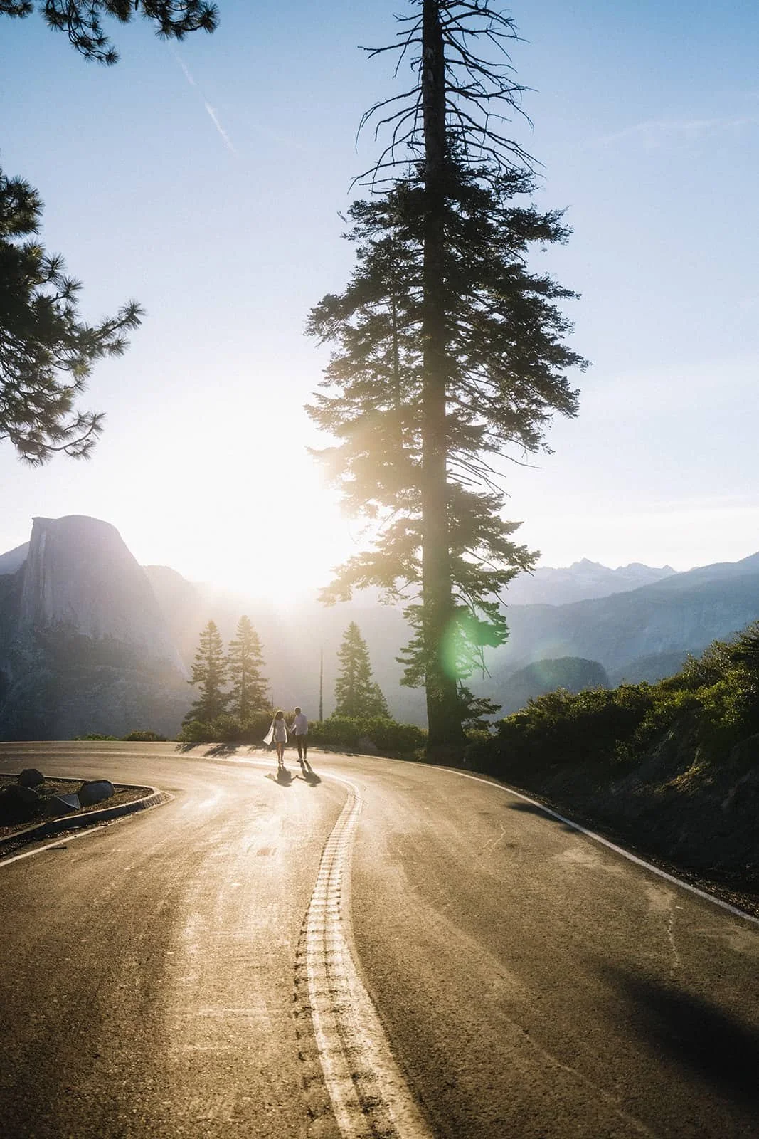 Couple walking hand in hand along a winding mountain road at Glacier Point in Yosemite during their engagement session, tall pines and distant mountains behind them