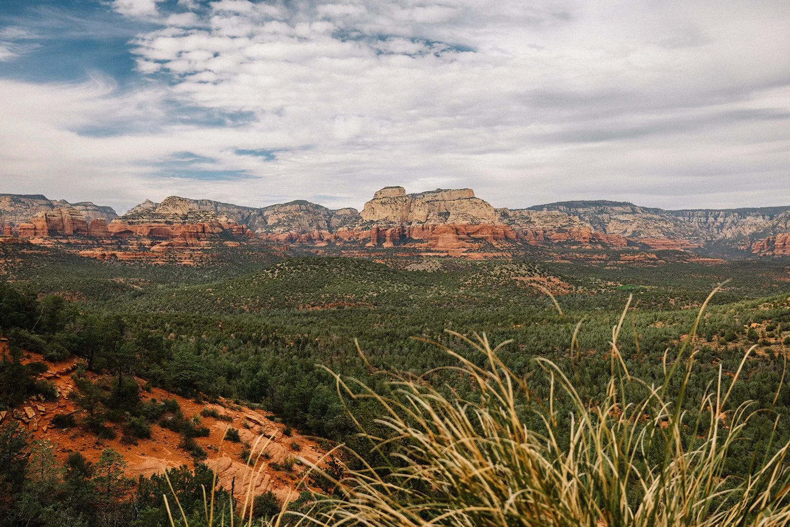 Landscape view of Sedona's red rock formations with green desert vegetation in the foreground and partly cloudy sky.