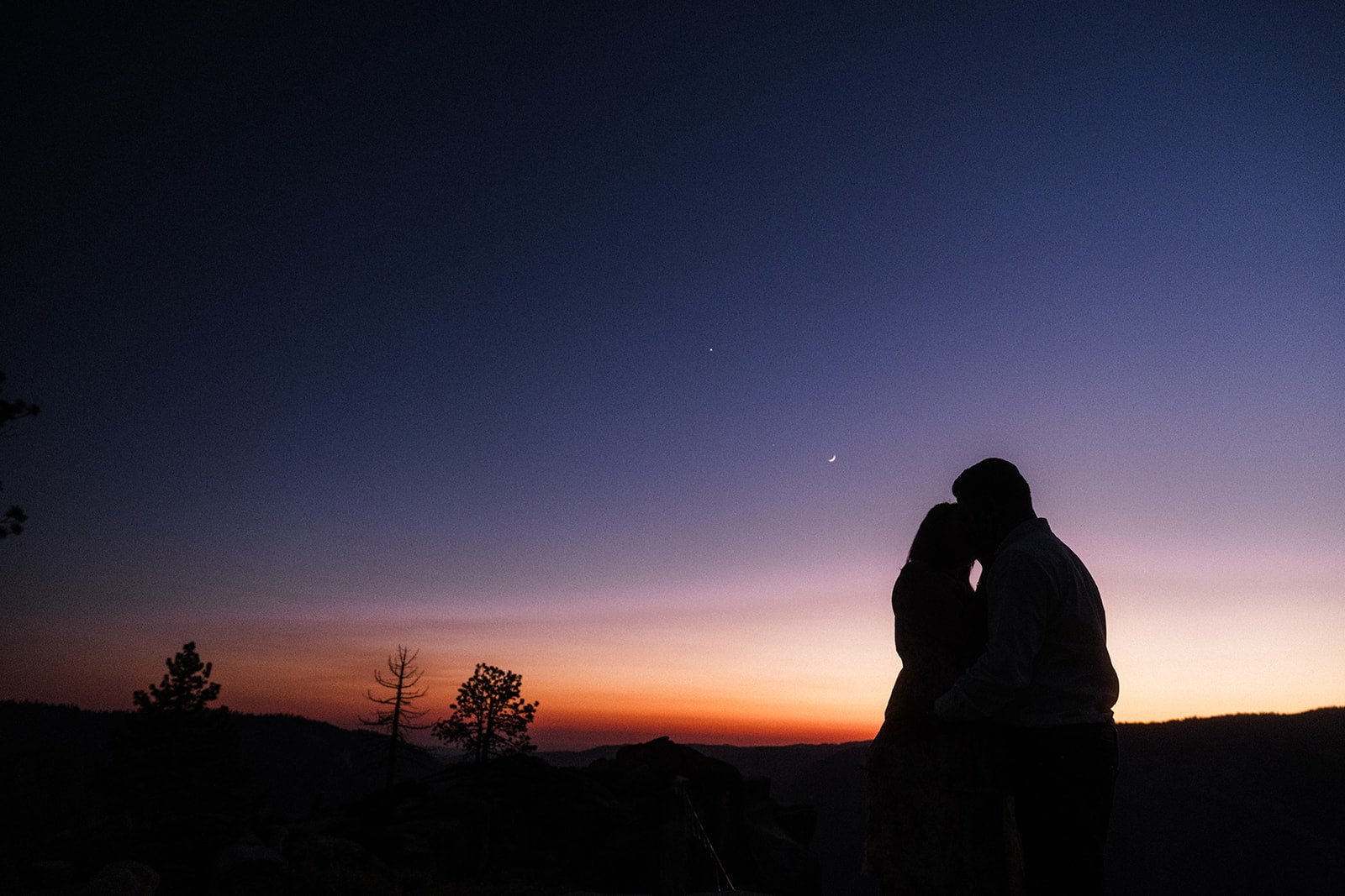Silhouette of a couple kissing at Taft Point in Yosemite at sunset during their engagement session, colorful sky with a crescent moon and stars