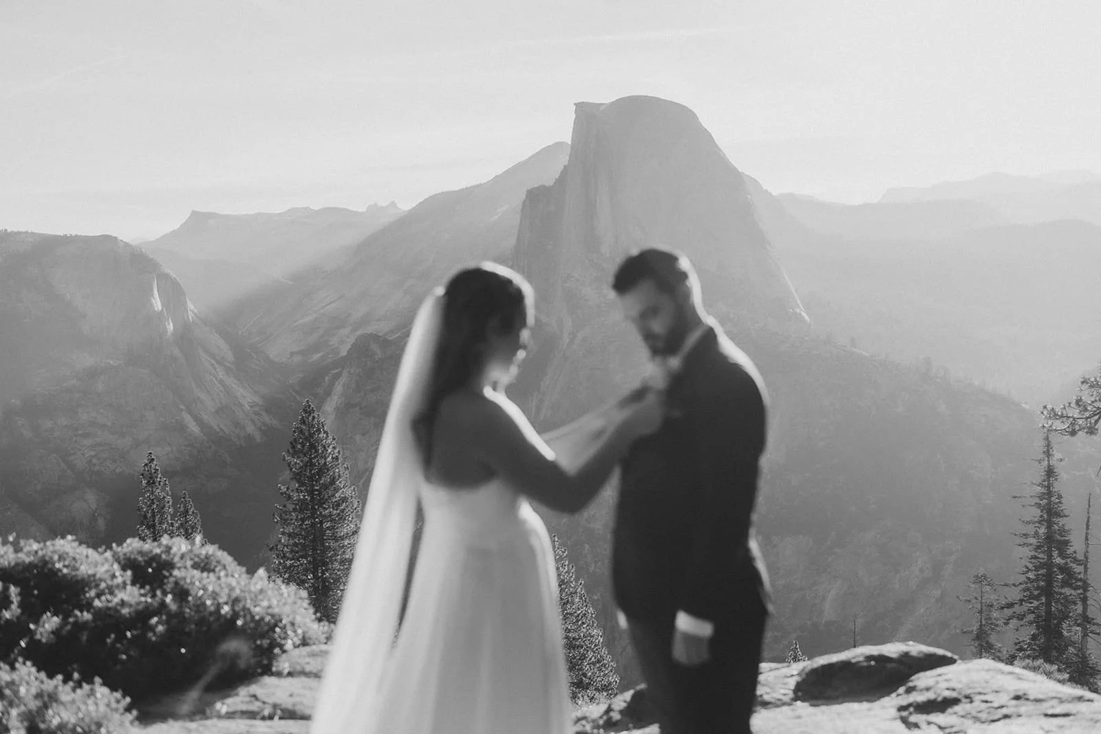 A black and white photo of a bride and groom holding hands and looking at each other outdoors with a mountain range background.