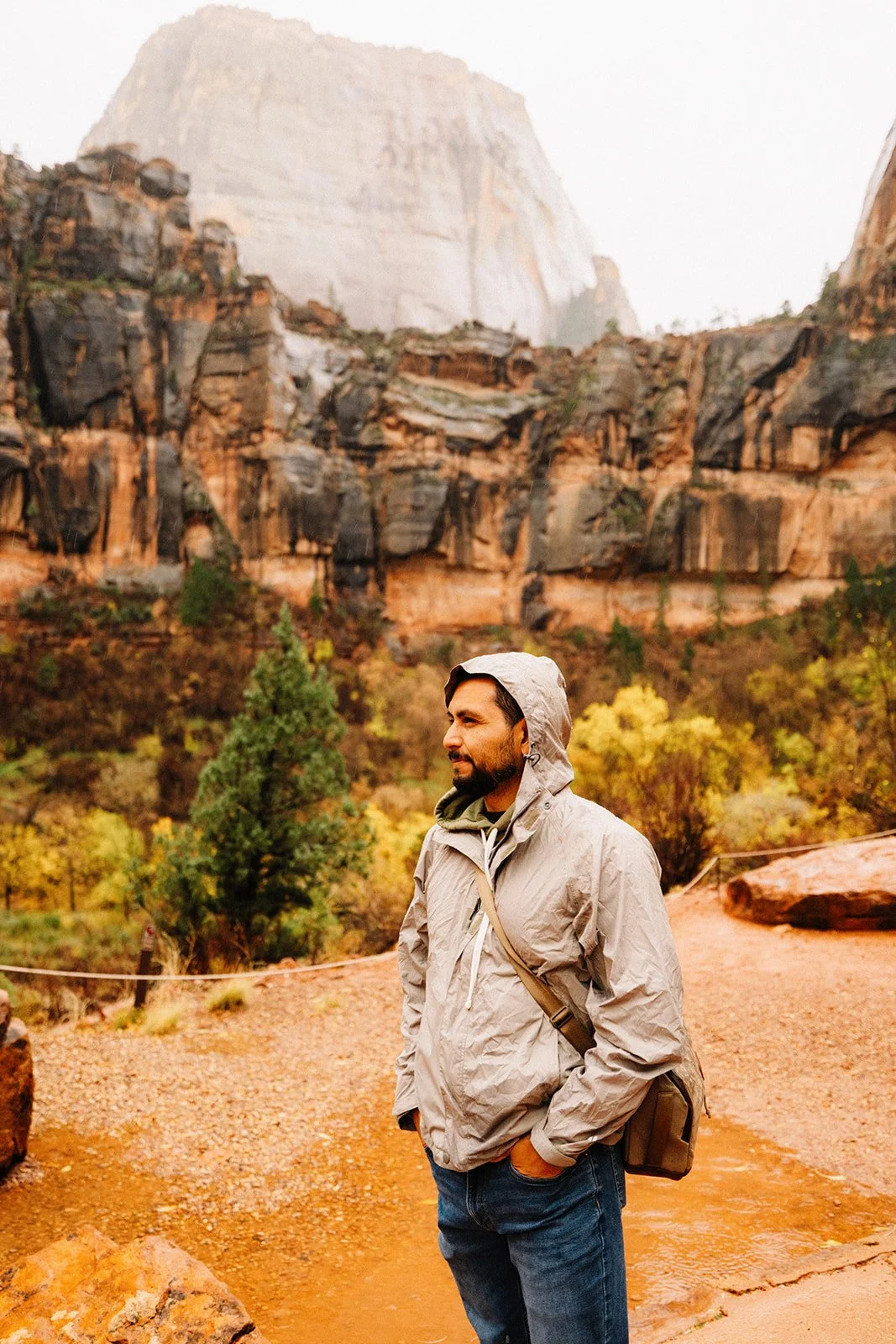 Armando, wearing a gray jacket and carrying a brown bag stands outside in front of colorful autumn trees and rocky cliffs, with a large mountain in the background.