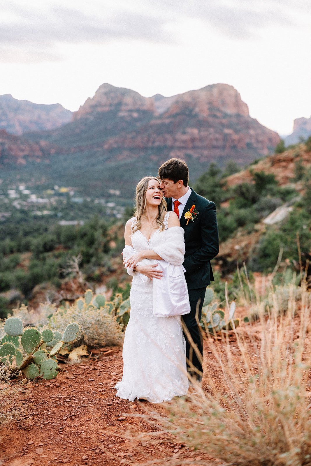 A newlywed couple stands on a dirt path in a desert landscape with cacti and shrubs, with red mountains in the background. The bride is wearing a white lace wedding gown and the groom is in a dark suit with a boutonniere. They are smiling and sharing an intimate moment.