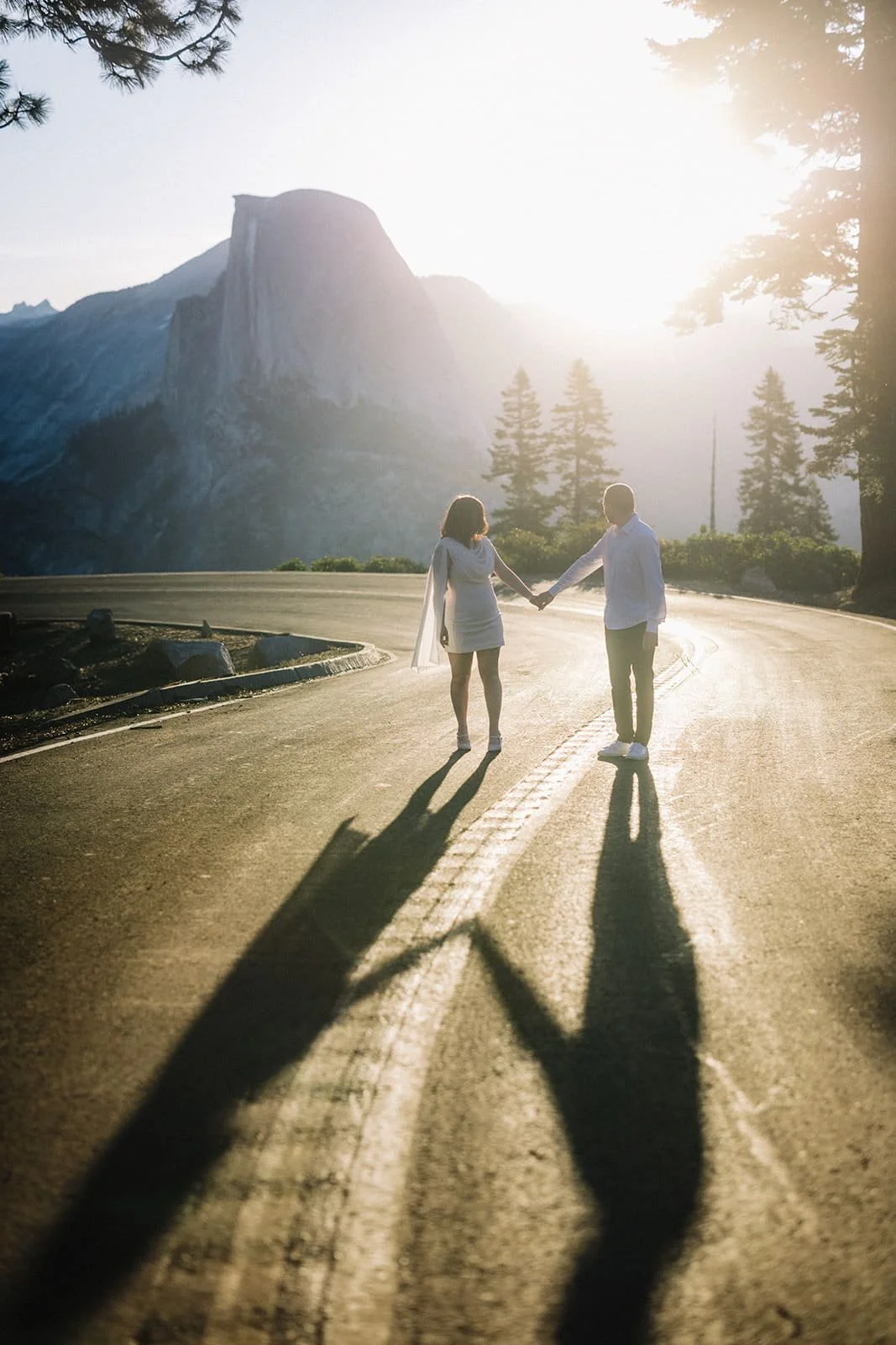 Couple holding hands on a winding mountain road at Glacier Point in Yosemite, sun setting behind the mountains and casting long shadows