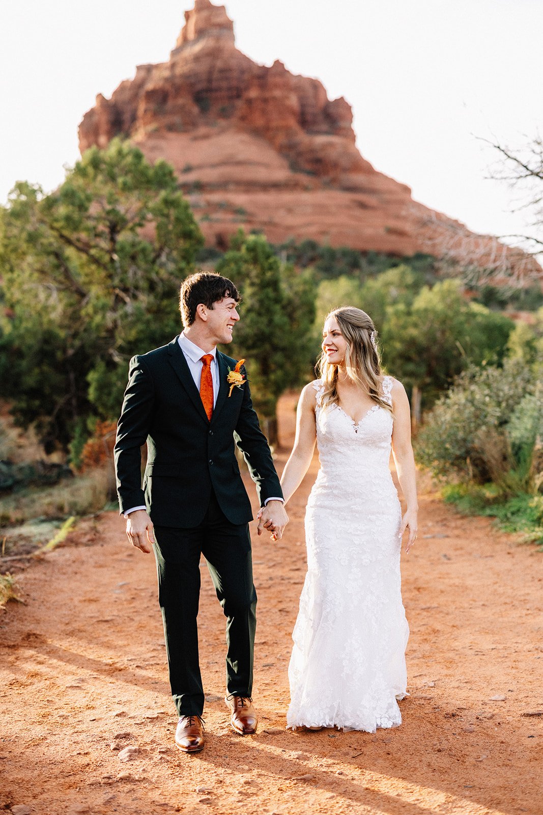 A couple in wedding attire walking on a dirt path with red rock formations and green trees in the background.