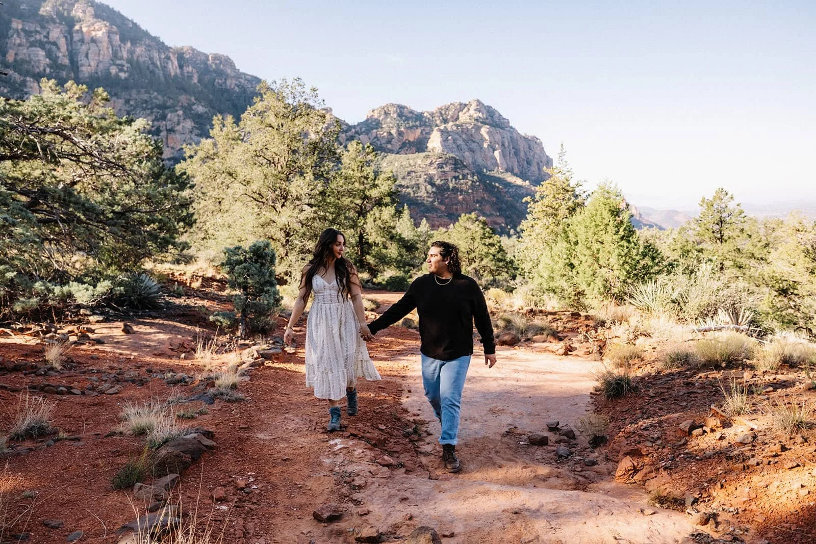 Couple holding hands walking a red rock trail during their Sedona engagement session, mountains and desert trees in the background