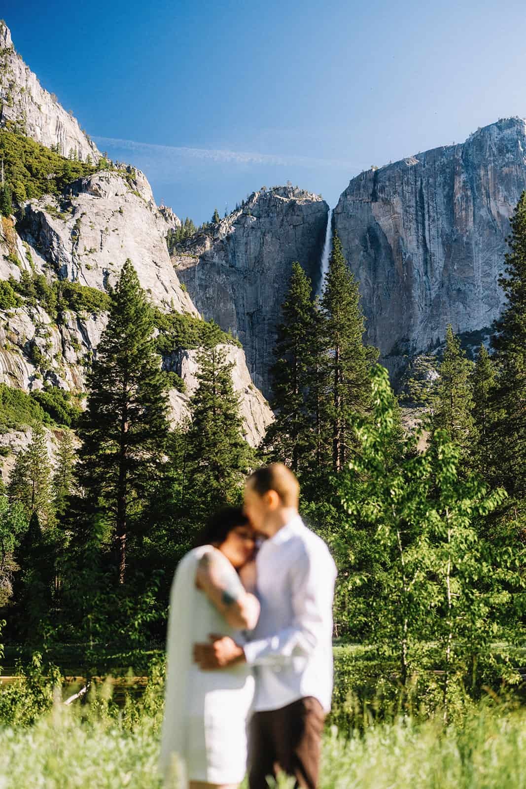 Couple standing close together at Cooke's Meadow in Yosemite, tall pines and granite cliffs behind them with a waterfall under a clear blue sky