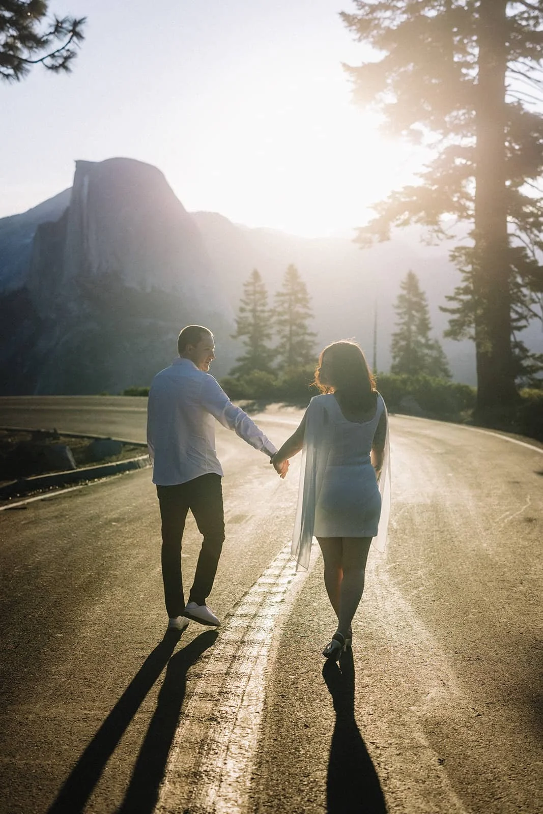 Couple holding hands walking along an empty road at Glacier Point in Yosemite at sunset, mountains and trees in the background