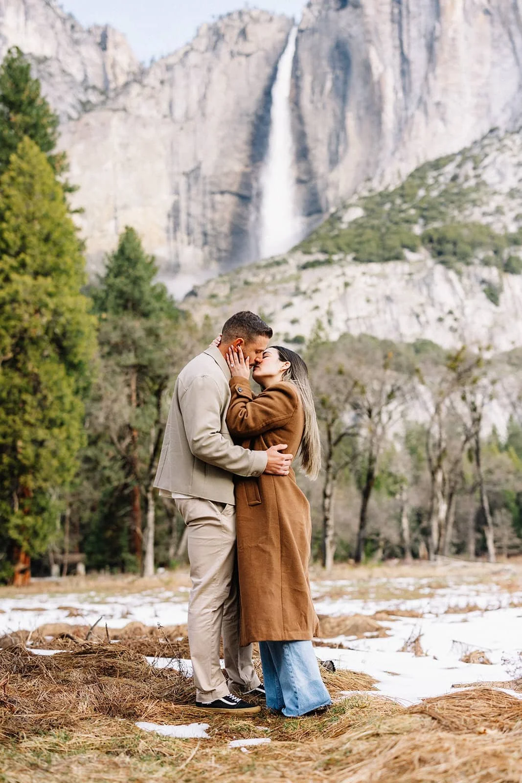 Couple embracing at Cooke's Meadow in Yosemite during their engagement session, a waterfall visible in the background