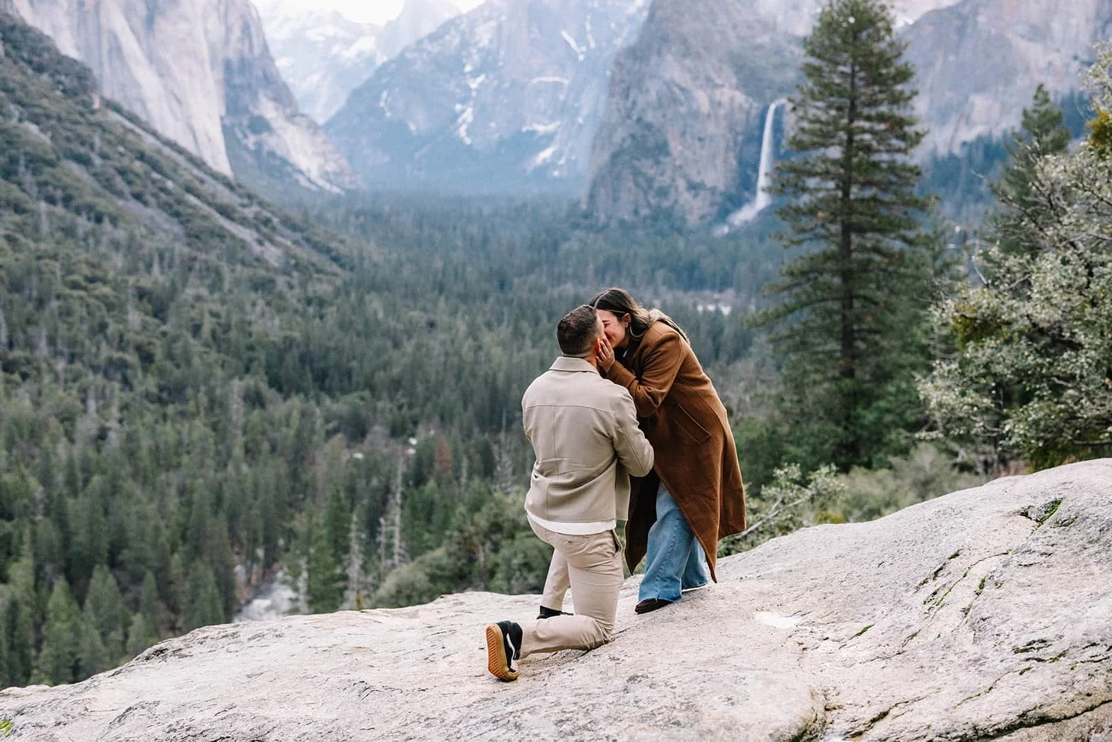 Man kneeling in proposal on a rock at Tunnel View in Yosemite, forested mountain landscape and waterfall in the background
