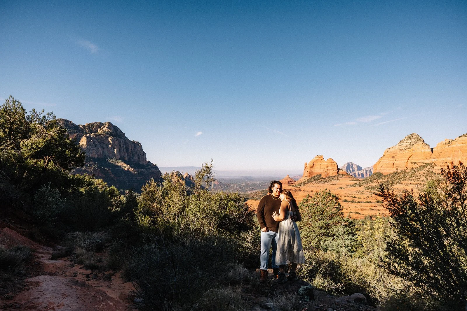 A couple standing close together on a trail in a desert landscape with red rock formations and sparse vegetation, under a clear blue sky.
