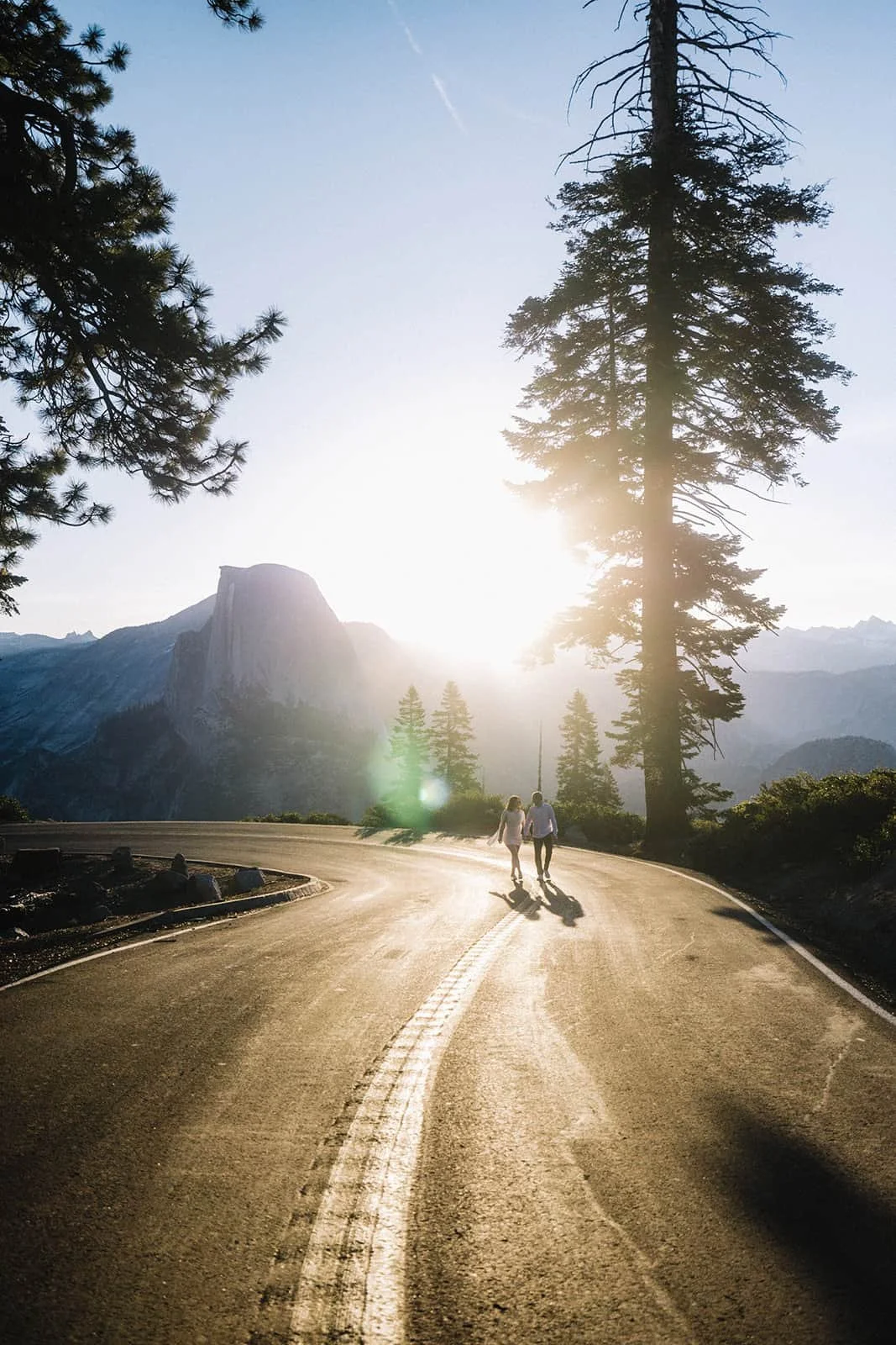 Couple walking hand in hand along a winding mountain road at Glacier Point in Yosemite at sunset, tall pines lining the path