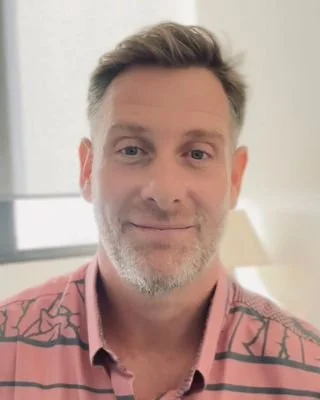 A middle-aged man with short hair and a beard, smiling, wearing a pink shirt with black geometric patterns, sitting indoors with a window and a lamp in the background.