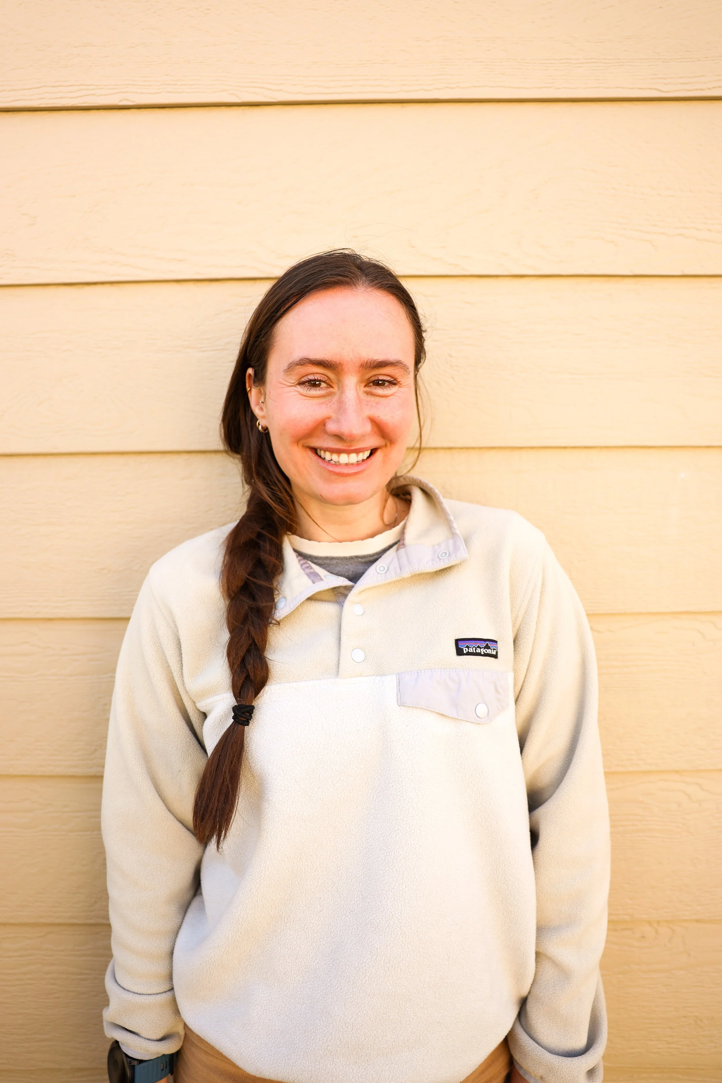 A smiling woman with long brown hair in a braid, wearing a beige fleece jacket, standing against a yellow wooden wall.