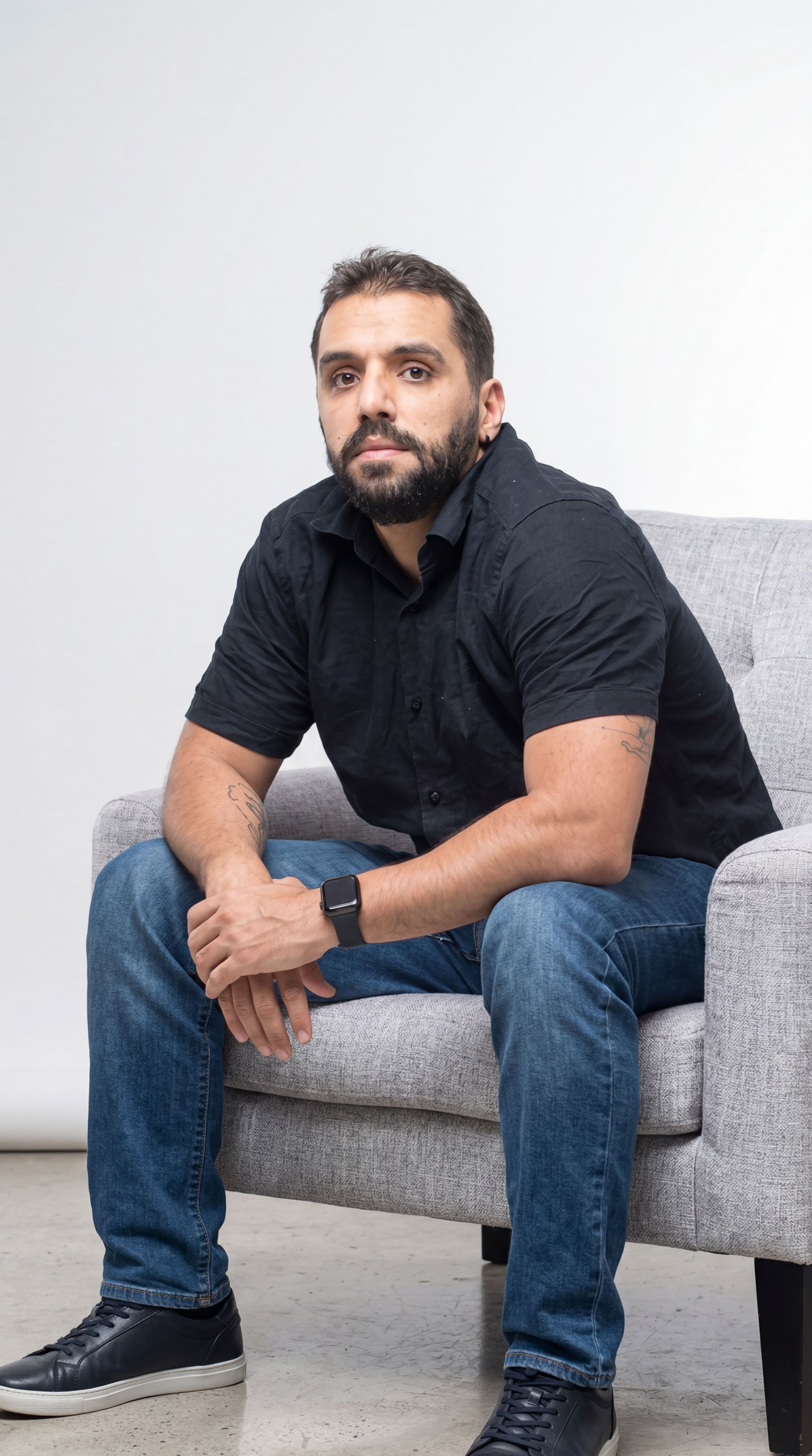 image of a man with a black button shirt and jeans seating on w grey seat resting his hand over his legs while looking at camera