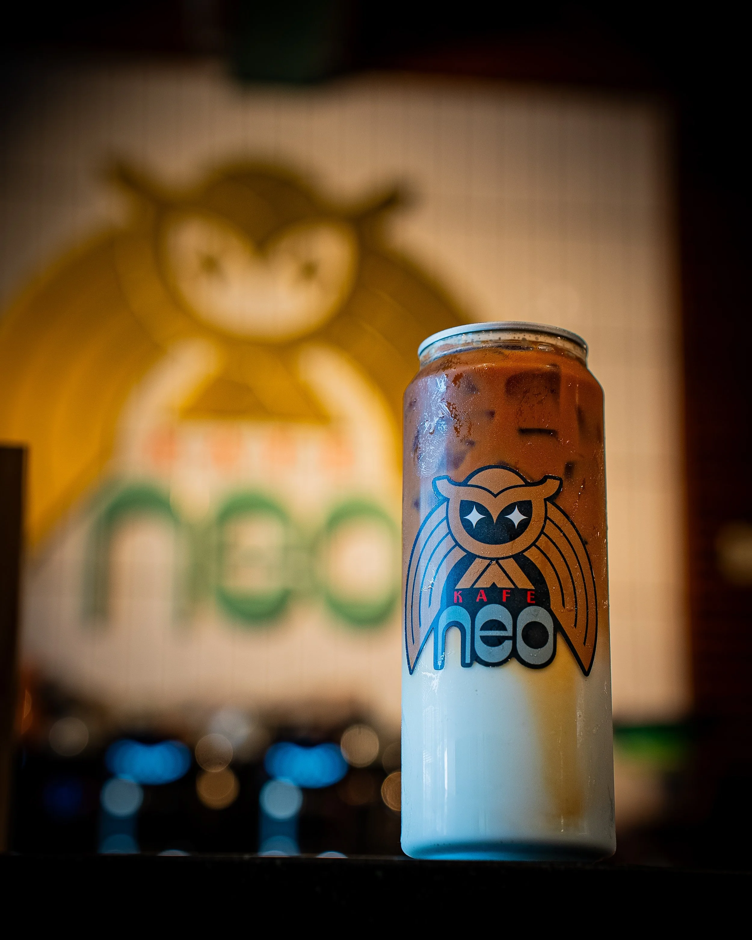 A can of iced coffee with a cartoon owl logo on it, set against a background of a blurry clock and colorful bokeh lights.