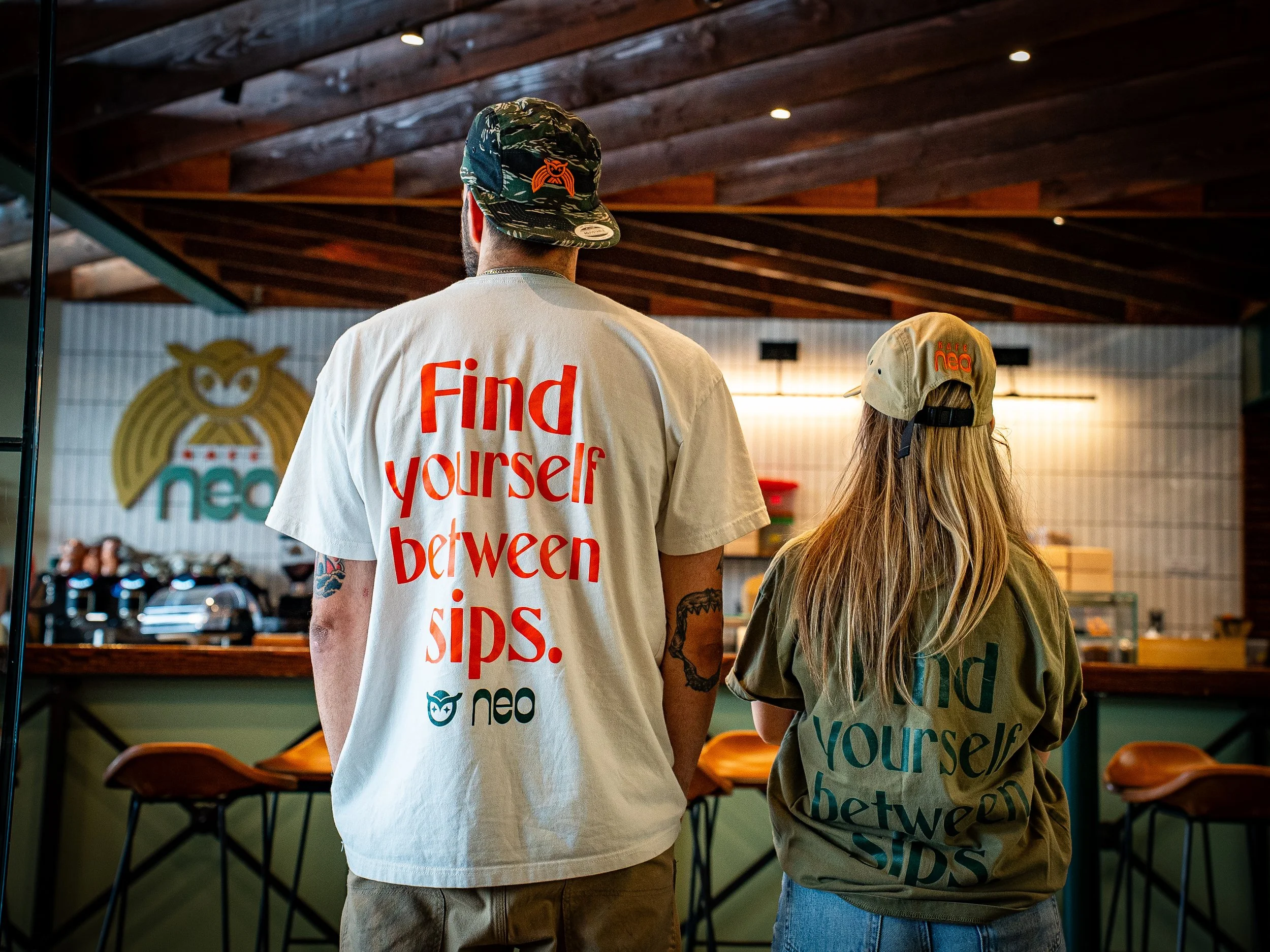 Two people, one man and one woman, stand inside a cafe or restaurant. The man wears a camouflage cap with an owl emblem and a white t-shirt with red and black text. The woman has long blond hair, a tan cap with the logo 'neo', and a green or gray shi