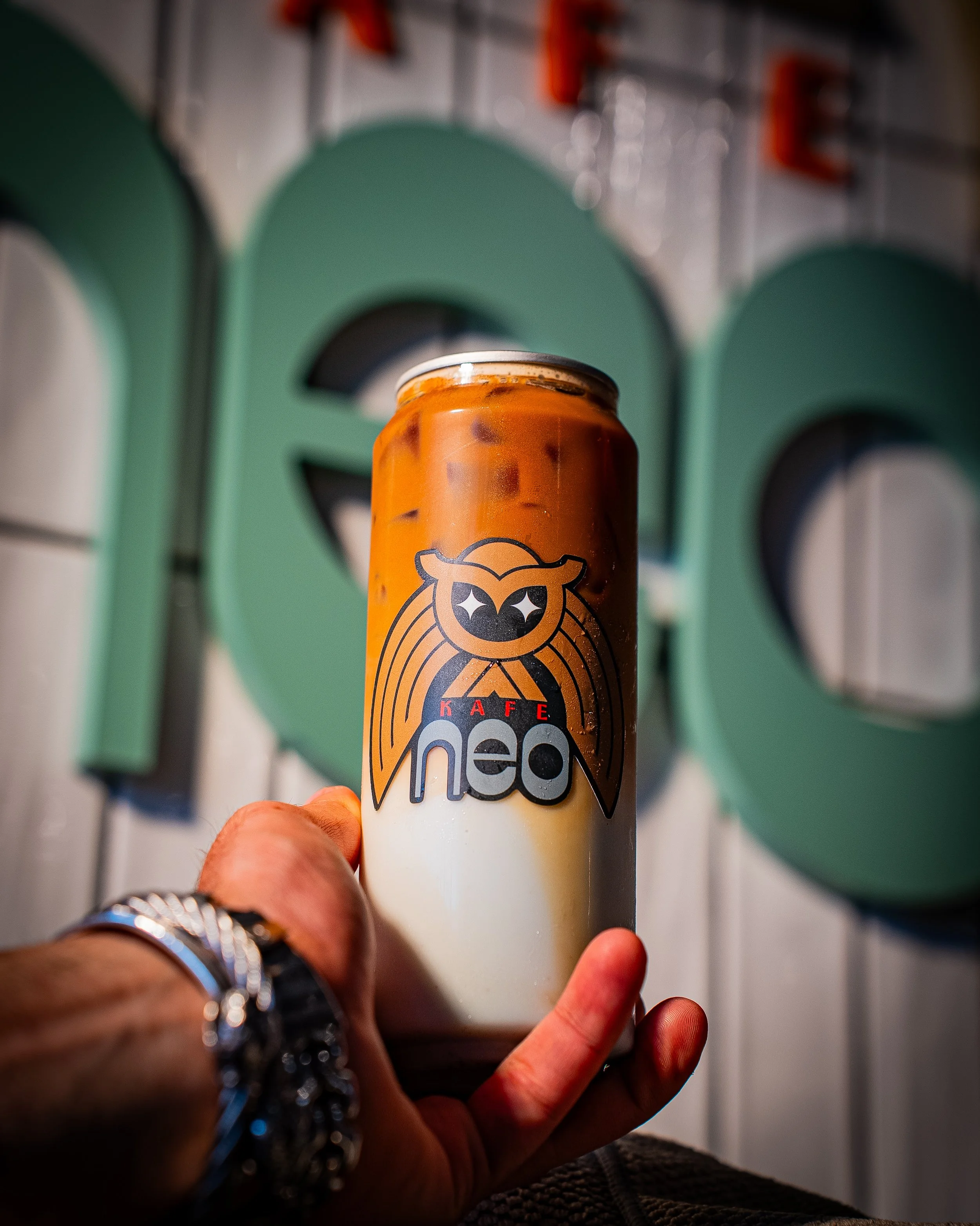 Close-up of a hand holding a can of coffee, with a logo of a smiling bear on it, in front of a colorful background that appears to be a vending machine or similar electronic display.