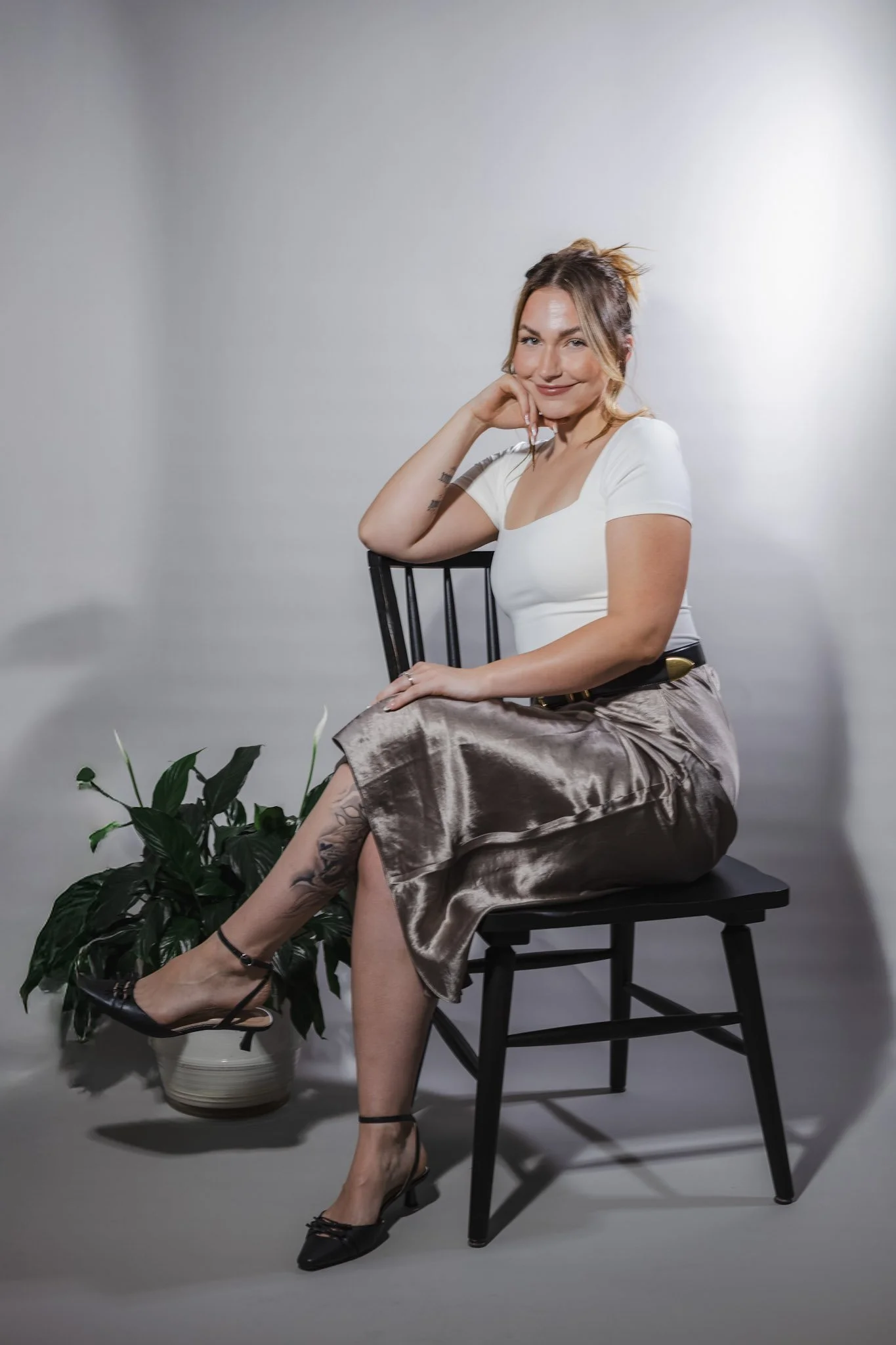 A woman sitting on a wooden chair, smiling, with her chin resting on her hand, wearing a white top, shiny satin skirt, black high heels, and has a tattoo on her ankle, next to a potted green plant.