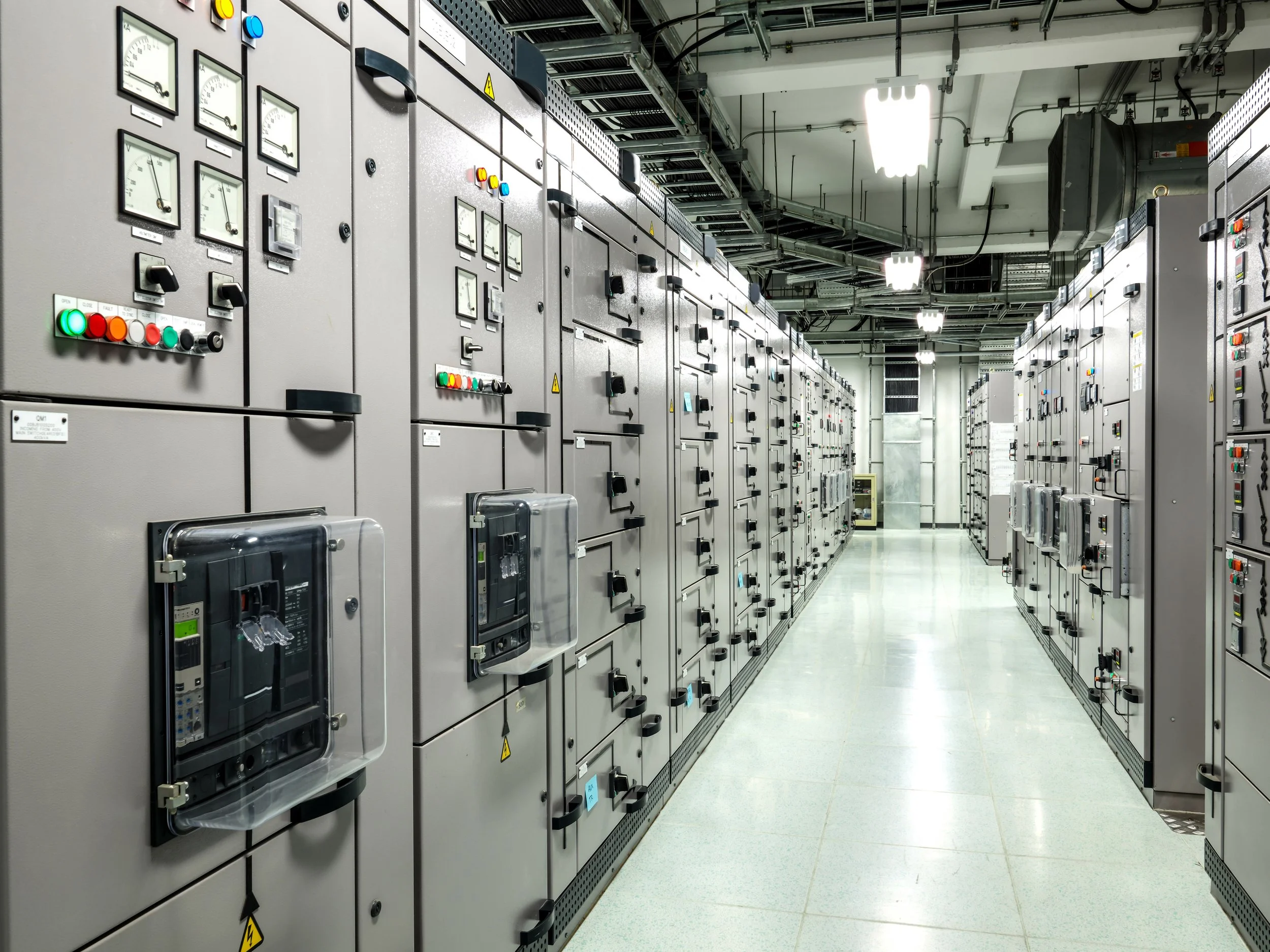 An industrial control room with rows of large gray electrical control panels with various switches, meters, and indicator lights.