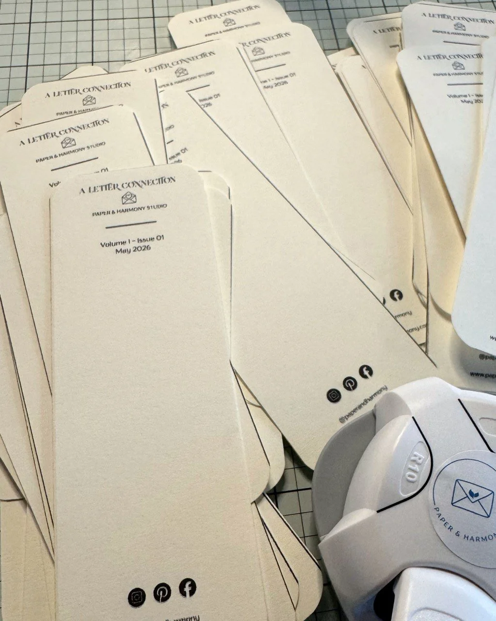 Several cream-colored bookmarks with black text and icons for social media platforms placed on a work surface, along with a paper puncher, all for a publication called "A Letter Connection" from Paper & Harmony Studio.