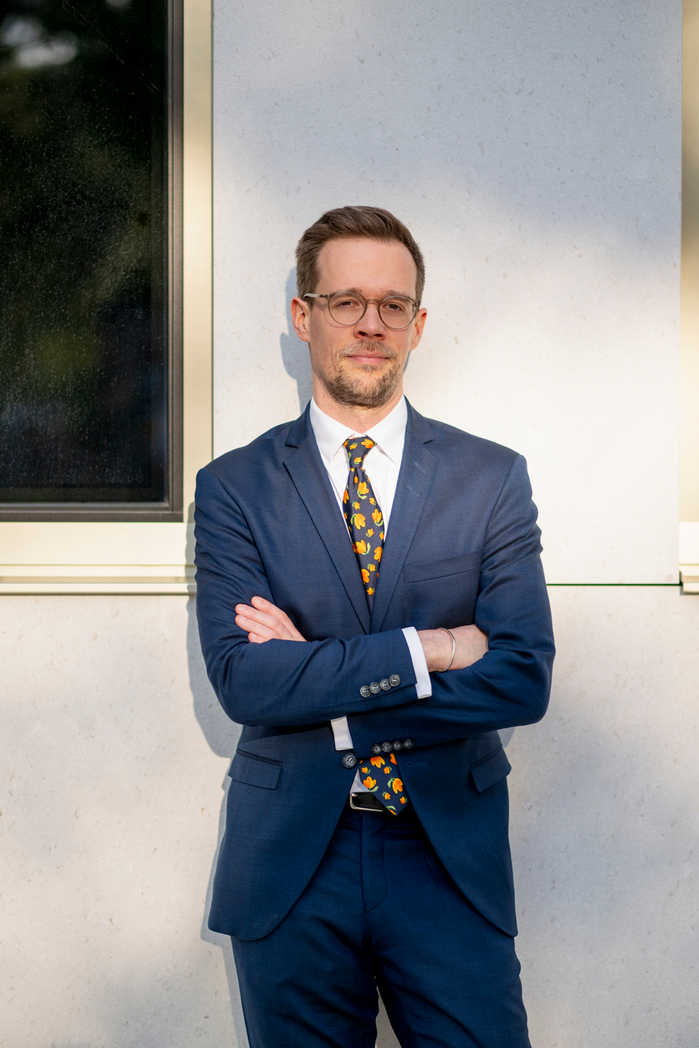 Rechtsanwalt Christoph Trafoier im blauen Anzug, weißem Hemd und einer bunten Krawatte. Er steht im Sonnenlicht vor einer weißen Wand.