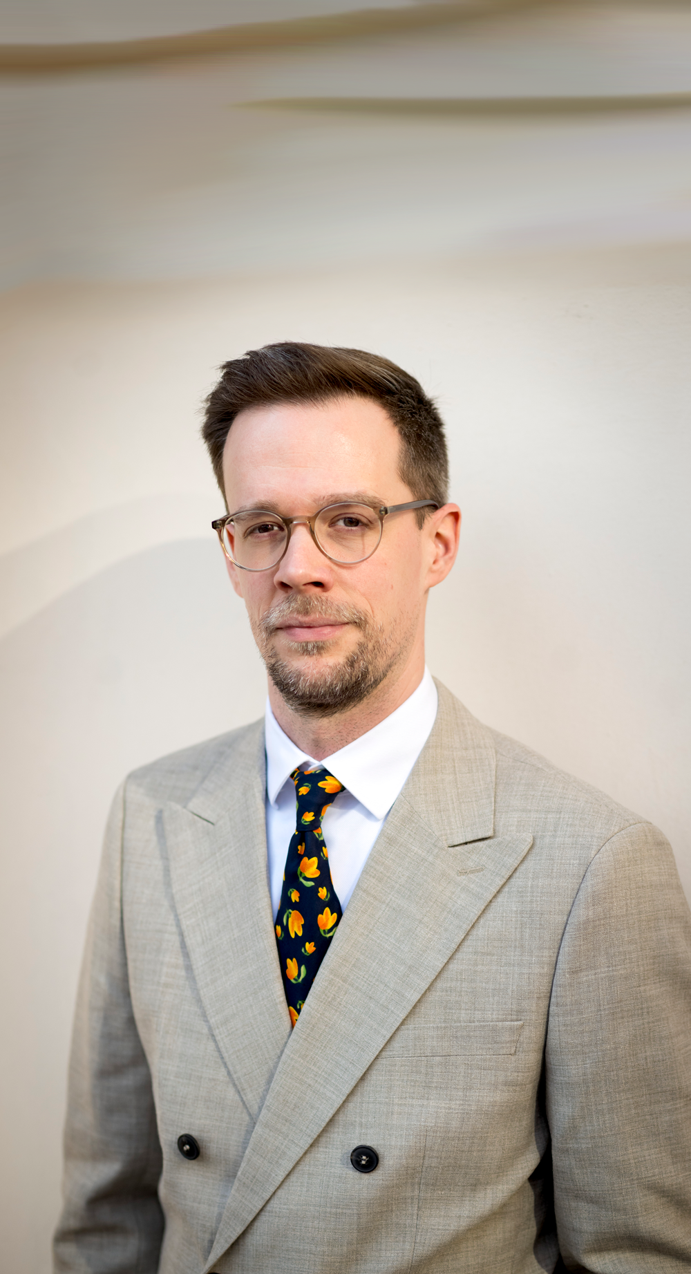 Rechtsanwalt Christoph Trafoier im grauen Anzug mit weißem Hemd und einer Krawatte mit orangen Blumen. Er steht vor einem neutralen Hintergrund.