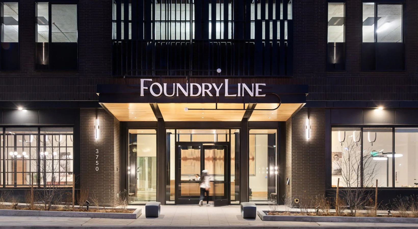 Night view the entrance of the FoundryLine. A modern building with a dark facade, large windows, and a wooden entrance canopy with a sign reading "FoundryLine."
