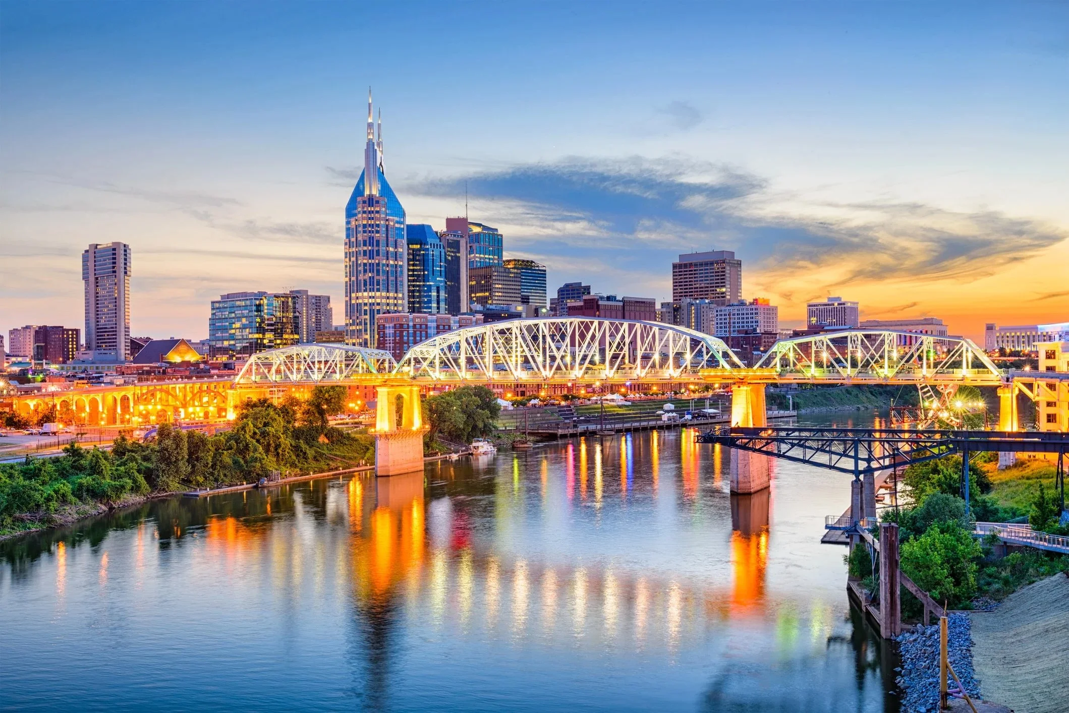 Nashville city skyline at sunset with tall skyscrapers, a river in the foreground reflecting the city lights, and a bridge crossing the river.
