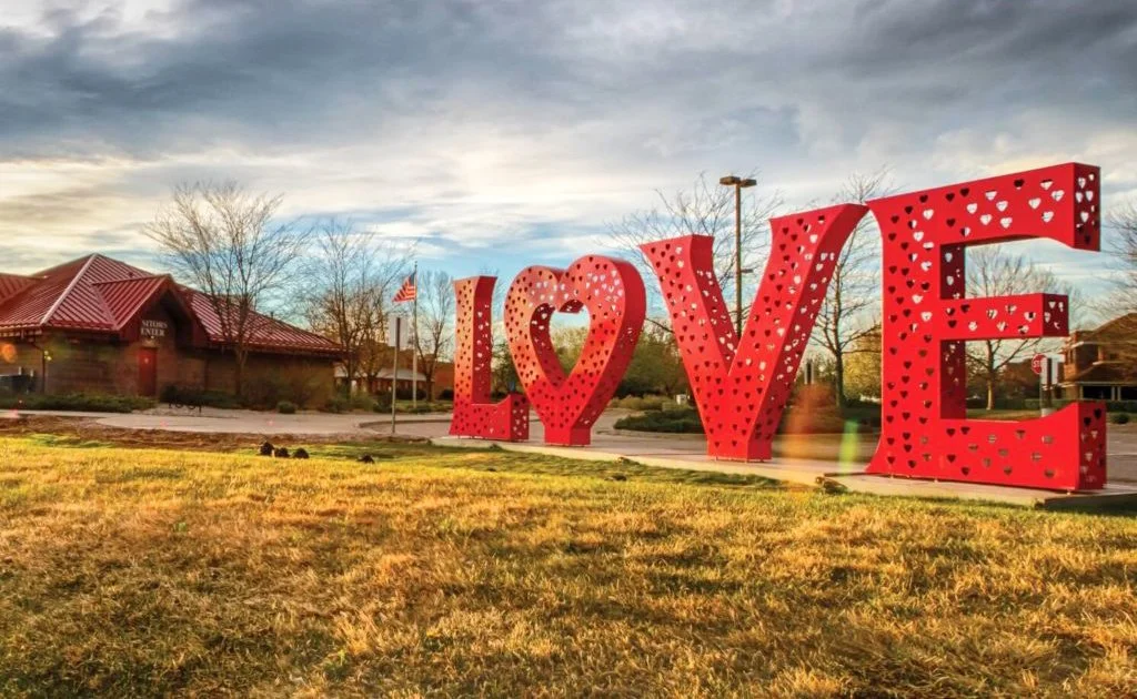 A large red sculpture spelling out the word 'LOVE' with a heart shape in place of the letter 'O.' located in a grassy area.