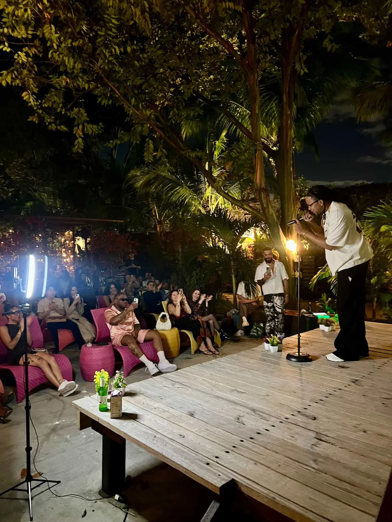 A performer singing into a microphone on stage at an outdoor event during nighttime, with an audience seated on colorful chairs and smiling.